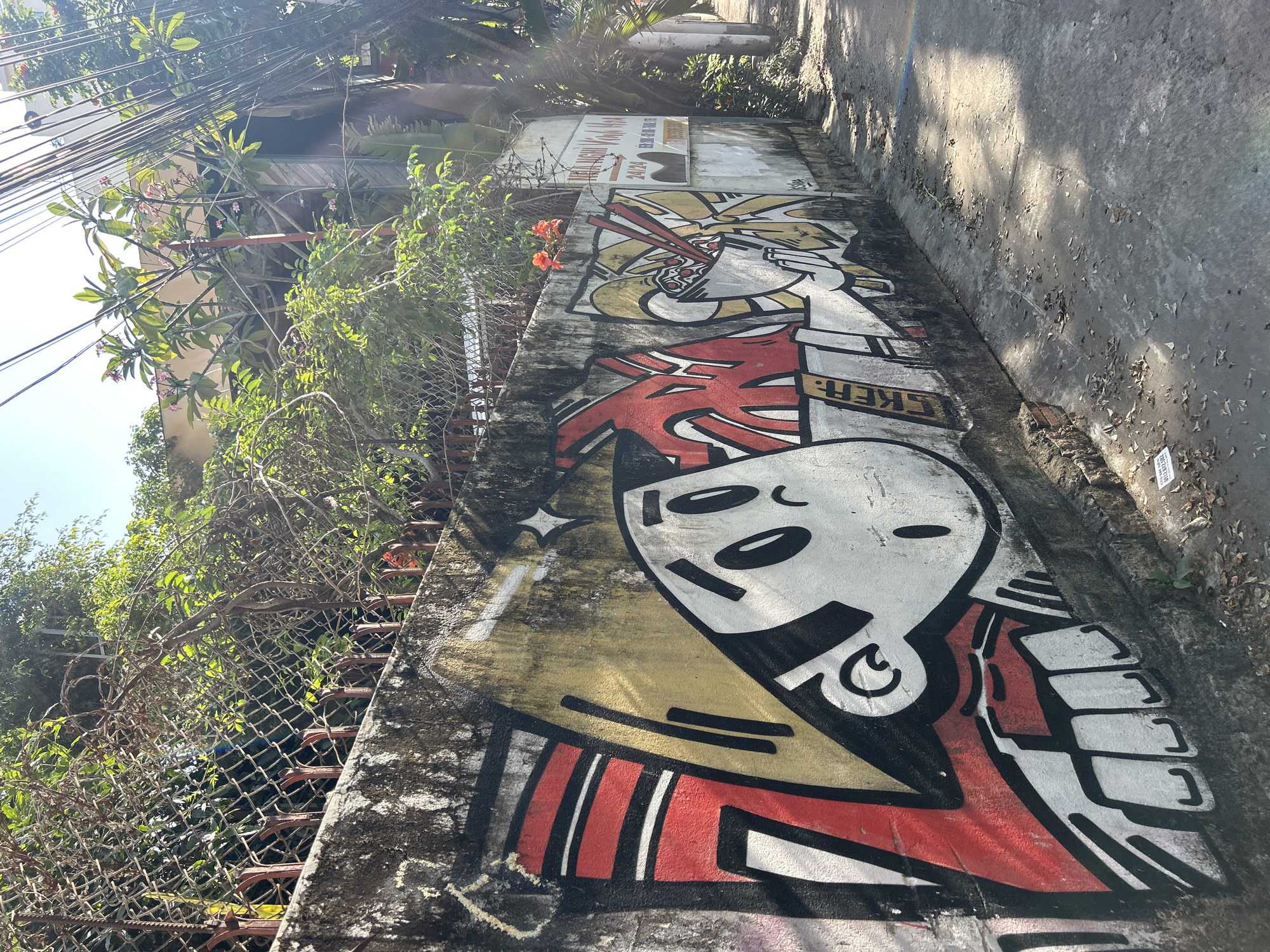 This media file presents a vibrant outdoor scene in Bắc Nha Trang, Vietnam, featuring a concrete wall covered in colorful street art. The wall borders a narrow, paved pathway, with lush tropical vegetation growing abundantly behind and above it.
The primary subject is a large, stylized graffiti mural. On the left, a cartoonish character with a large white head, black eyes, and a small, simple mouth is depicted. Its body is predominantly red, with distinct black and white block-like patterns, and yellow and black accents outline its head. The partial word "IKER" is visible in block letters on a white section of its arm or side. Further along the wall to the right, another section of graffiti portrays a bowl of noodles, likely ramen or phở, with chopsticks emerging from it, rendered in yellow, white, and red with black outlines. A star-shaped sparkle is painted near the main character's head.
Above the noodle graffiti, a light-colored sign is affixed to the wall, displaying "24/24" prominently, alongside partially visible Vietnamese text, including "NHÀ NGHĨ HOÀNG AN" (Hoang An Guesthouse/Hotel).
The surrounding environment is characterized by dense green foliage, including bushes and trees with pink frangipani-like flowers, and some orange-red blossoms. A chain-link fence with rebar supports is visible through the greenery on the left. Numerous black utility lines crisscross the bright, clear sky, indicating a sunny daytime setting. Sunlight casts distinct shadows on an adjacent, unpainted concrete wall. The ground next to the graffiti-covered wall is a concrete path scattered with fallen leaves.