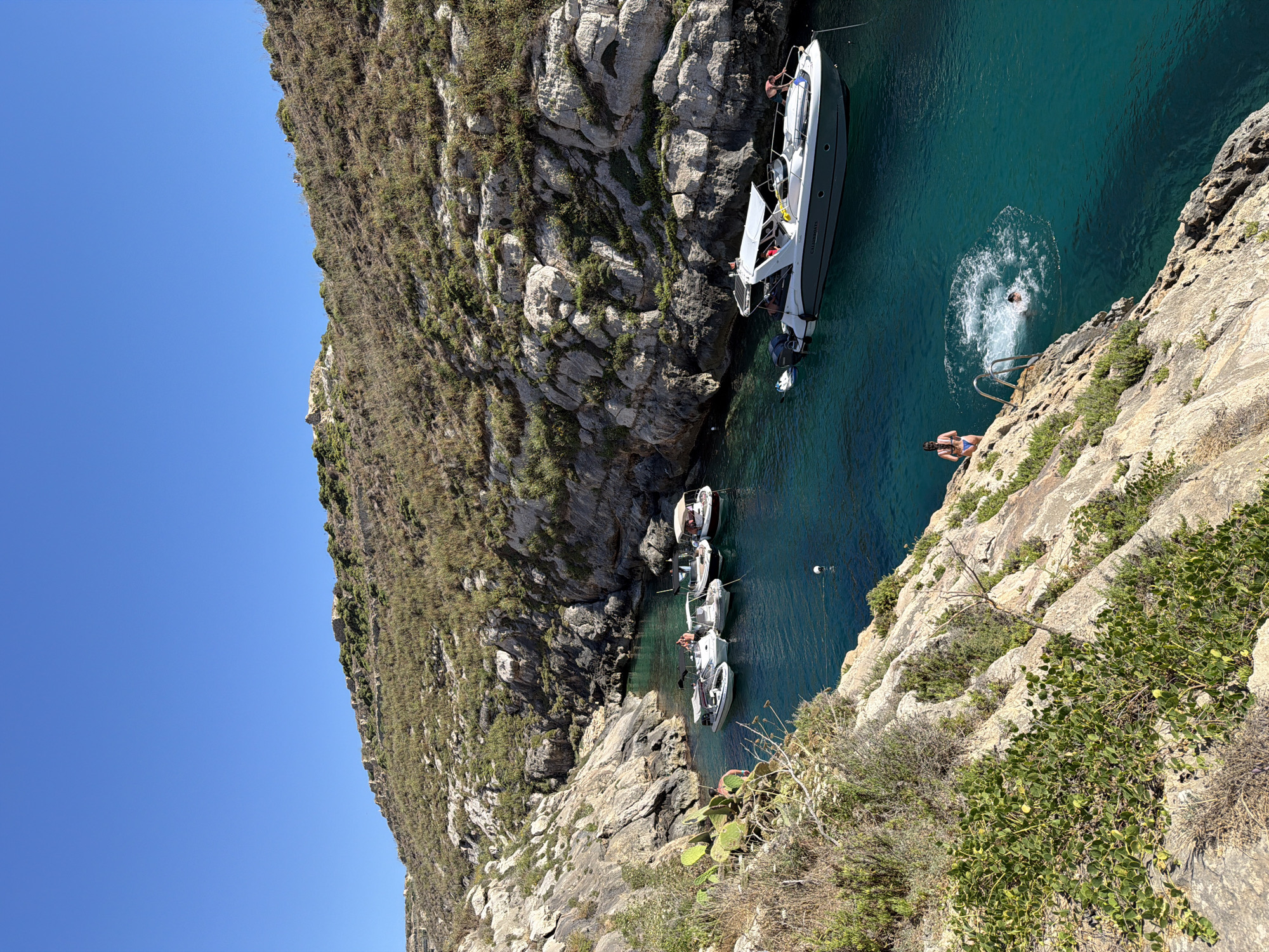 A high-angle, full outdoor shot depicts a small, rocky cove in Xlendi, Malta, under a clear blue sky. The water is a vibrant turquoise, revealing a rocky seabed. Several small boats are moored in the cove, with a larger white motorboat prominently positioned to the right. On the rocky shore in the foreground, a person in a bikini stands near a metal ladder that leads into the water. Another person is in the water, creating a splash as they swim or dive. The surrounding cliffs are rugged and covered with sparse vegetation.