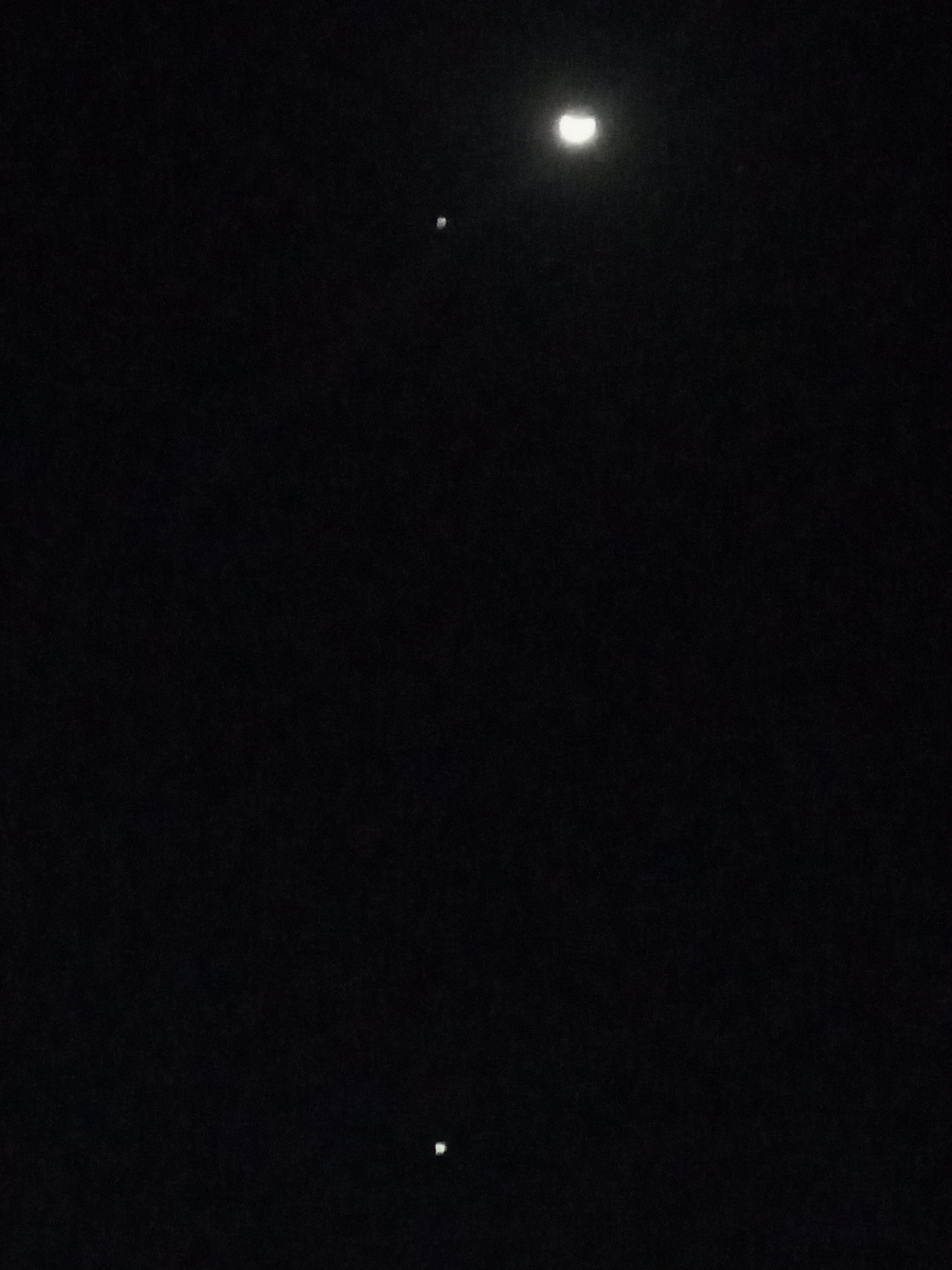The image shows a dark night sky. There is a crescent moon in the upper portion of the image and two point light sources positioned below it. The location of the image is said to be in Jos, Nigeria, though this cannot be confirmed from the visual evidence. There are no people or objects aside from the moon and the two lights.
