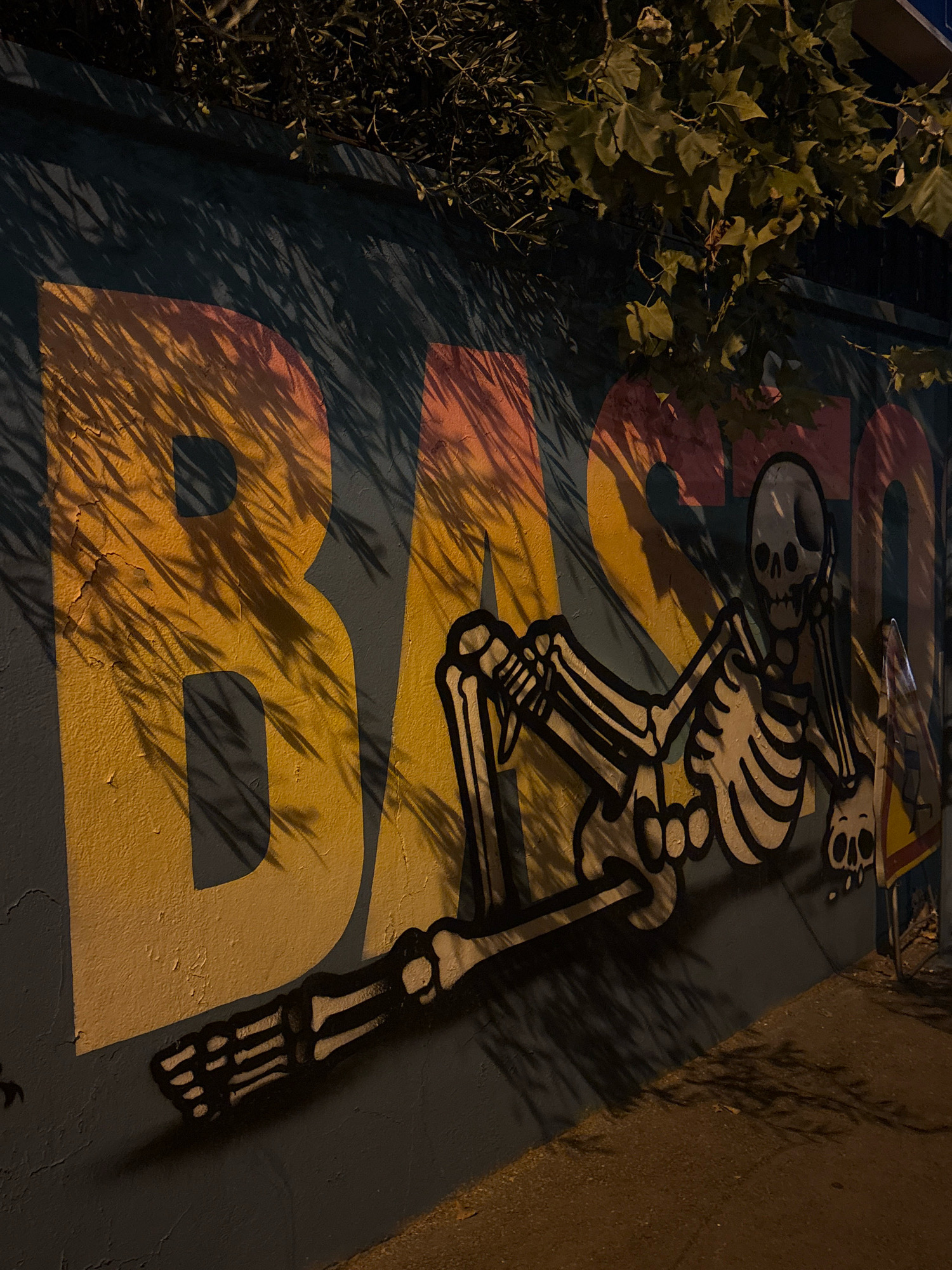 An outdoor mural on a dark blue wall features large, stylized letters in yellow and pink that read "BASSO". A cartoon skeleton is painted in black and white, appearing to lounge against the letters, with its arms reaching towards the "B" and its legs extending across the "S" and "O". The skeleton's head is positioned near the "O". Shadows from foliage are cast across the mural, obscuring parts of the letters and the skeleton. The ground in front of the mural is paved with a rough, brown surface. A small portion of a metal pole with a yellow and red sign is visible on the right edge of the image. The scene is illuminated by artificial light, creating a dramatic effect with the shadows.