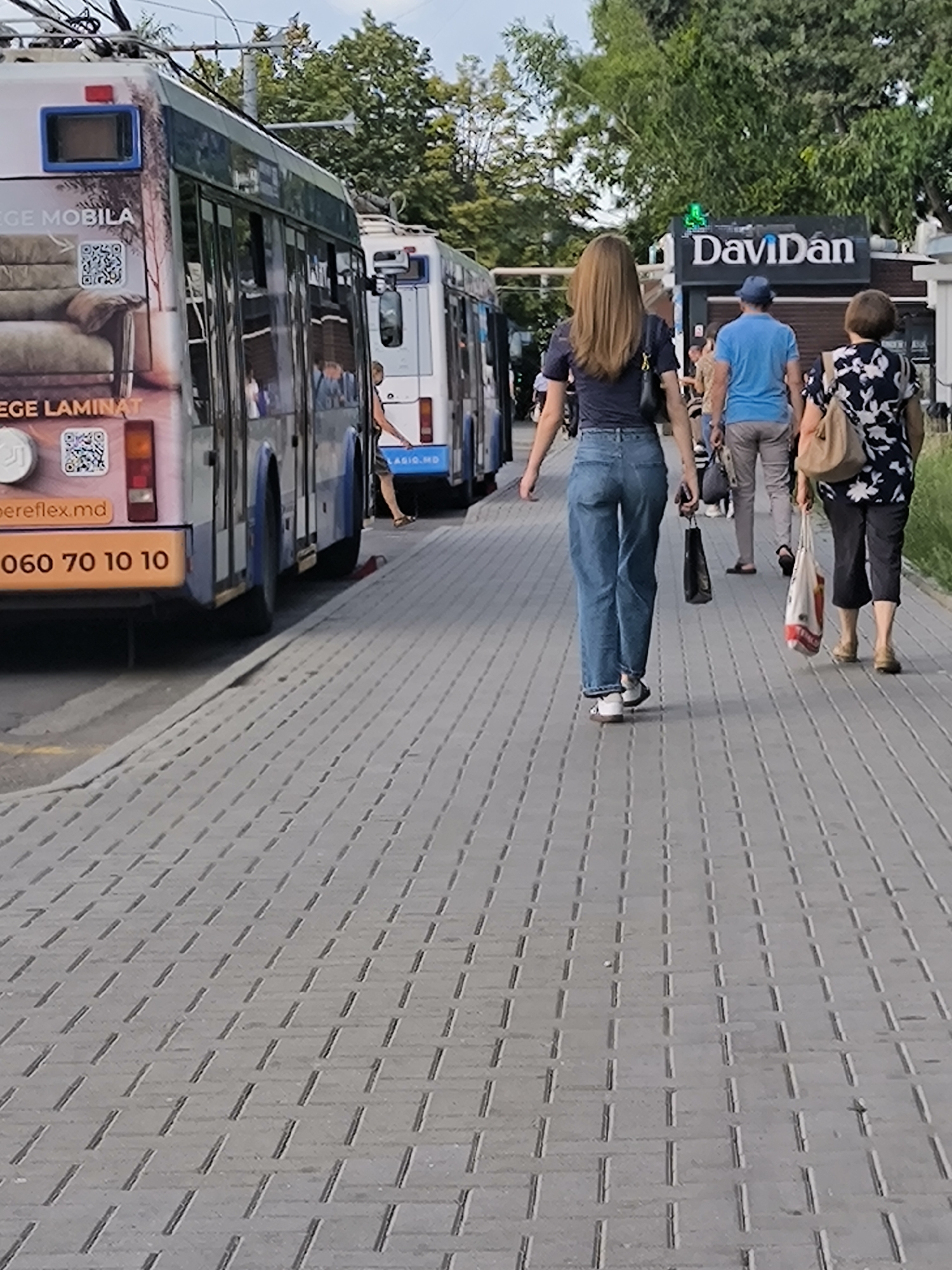 The image captures a street scene in Chișinău, Moldova, with a focus on a pedestrian walkway and public transportation. In the foreground, a woman with long brown hair walks away from the camera, wearing a dark blue t-shirt and light blue jeans. She carries a black handbag over her left shoulder. To her right, an older woman in a black and white floral dress walks towards the camera, carrying a patterned shopping bag in her right hand and a beige handbag over her left shoulder. Between them, a man in a blue t-shirt and grey trousers walks away from the camera, holding a dark bag in his right hand and wearing a blue hat. Behind them, a sign for "DaviDän" is visible.
On the left side of the image, a public bus, possibly a trolleybus due to the overhead wires, is parked or moving along the street. The bus has advertisements on its side, including text and QR codes. Another similar bus is visible behind the first one. The street is paved with grey interlocking bricks, and trees line the background, suggesting an urban environment with greenery. The lighting indicates it is daytime, likely late afternoon or early evening, given the shadows.
