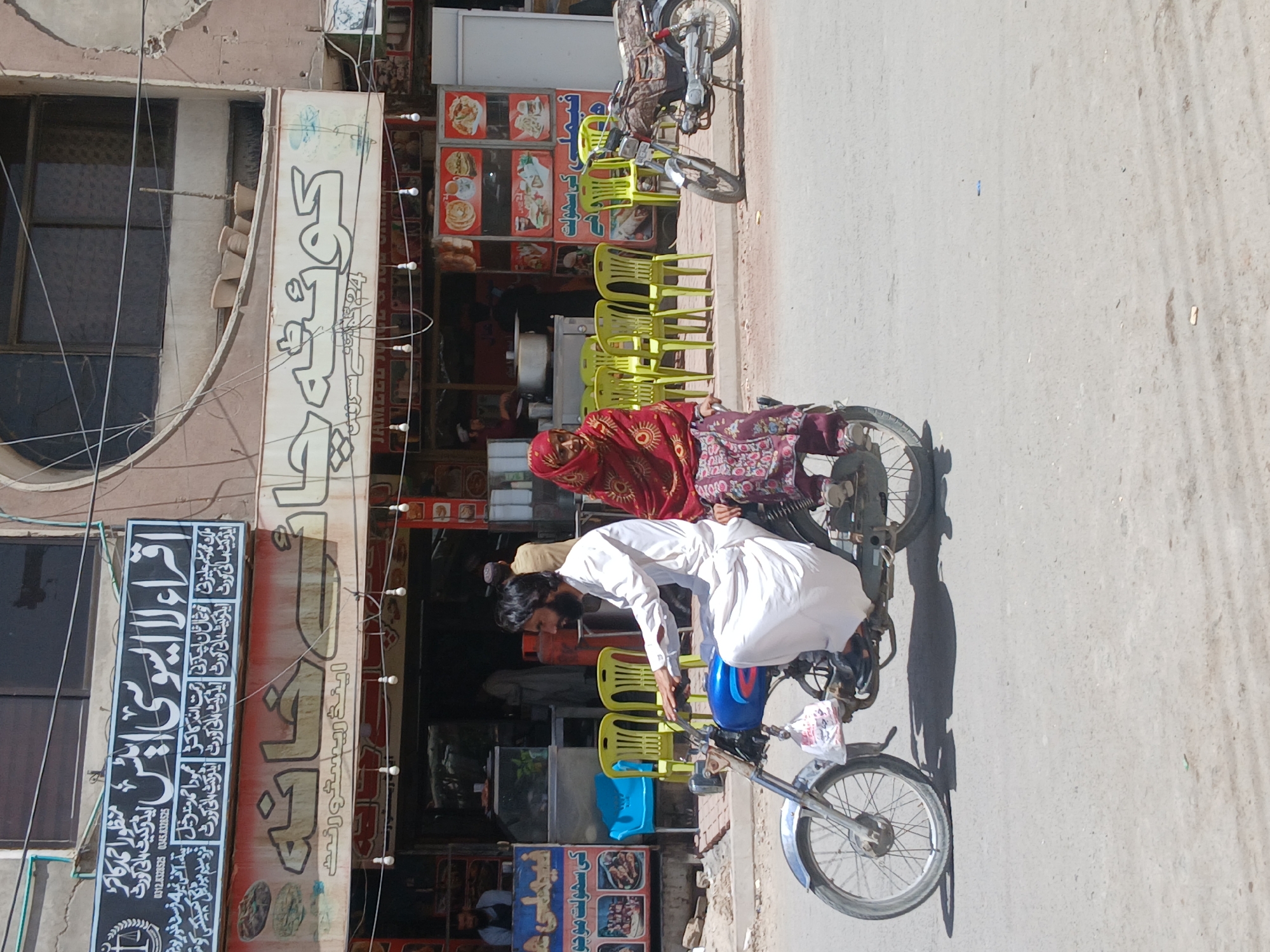 The image shows a man in a white shalwar kameez bending over a motorcycle in what appears to be a street in Quetta City Tehsil, Pakistan. A woman in a red headscarf and patterned clothing sits behind him on the motorcycle. The background includes a building with signs in Urdu, some with illuminated food menus, and stacks of green plastic chairs. There's another motorcycle parked beside the building. The scene suggests a commercial area, potentially a restaurant or food stall.