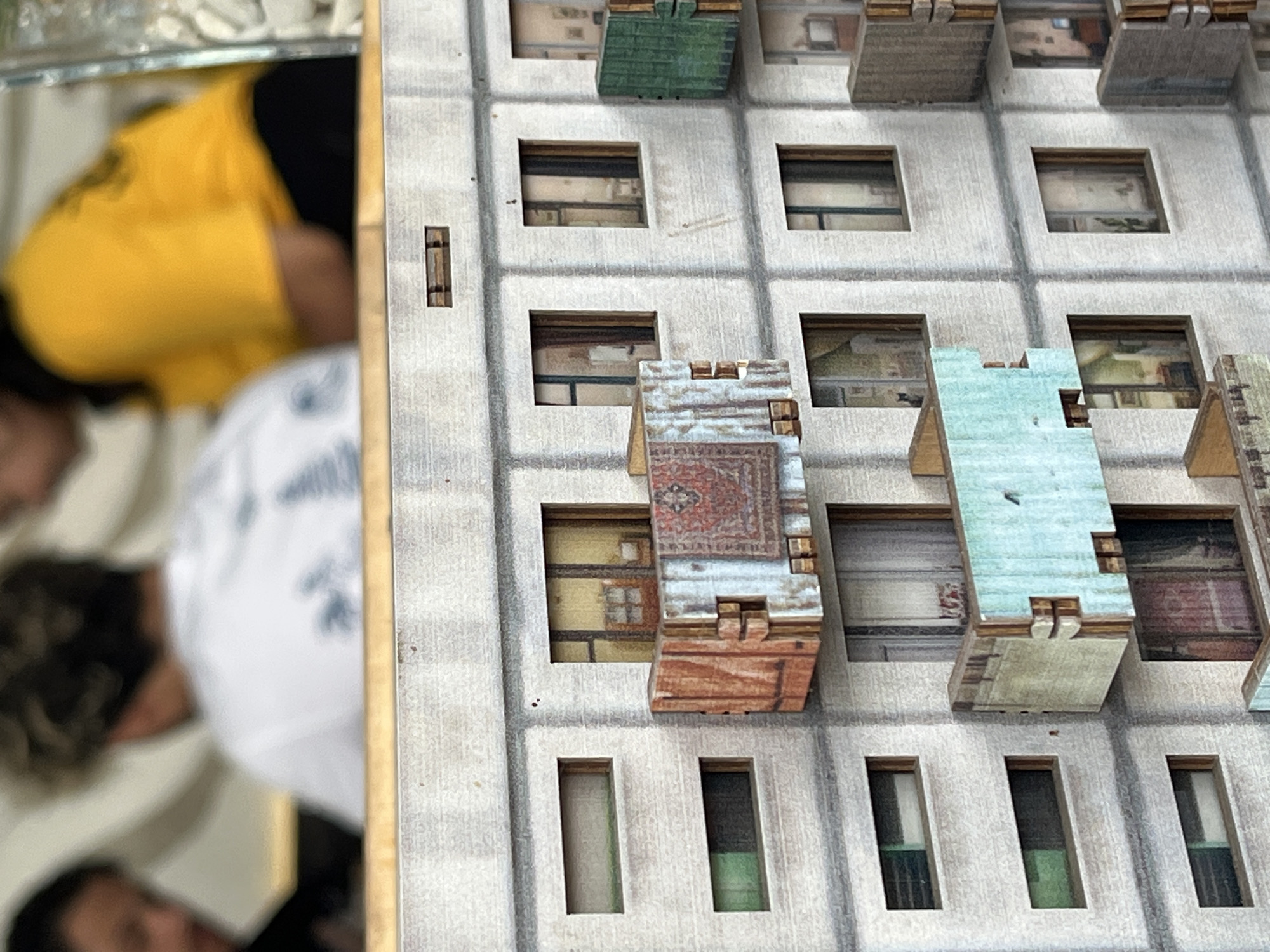 A close-up shot reveals a segment of a miniature model building with various window designs and facade textures. The building's architecture features repetitive window patterns, some with balconies or overhangs. The window interiors show different designs and colors. To the left of the model is a blurred reflection of people, including someone in a yellow shirt. Due to the lack of context in the image, determining the location of the building within Kuta Selatan, Indonesia, or any specific details about people, actions, or environment is impossible.