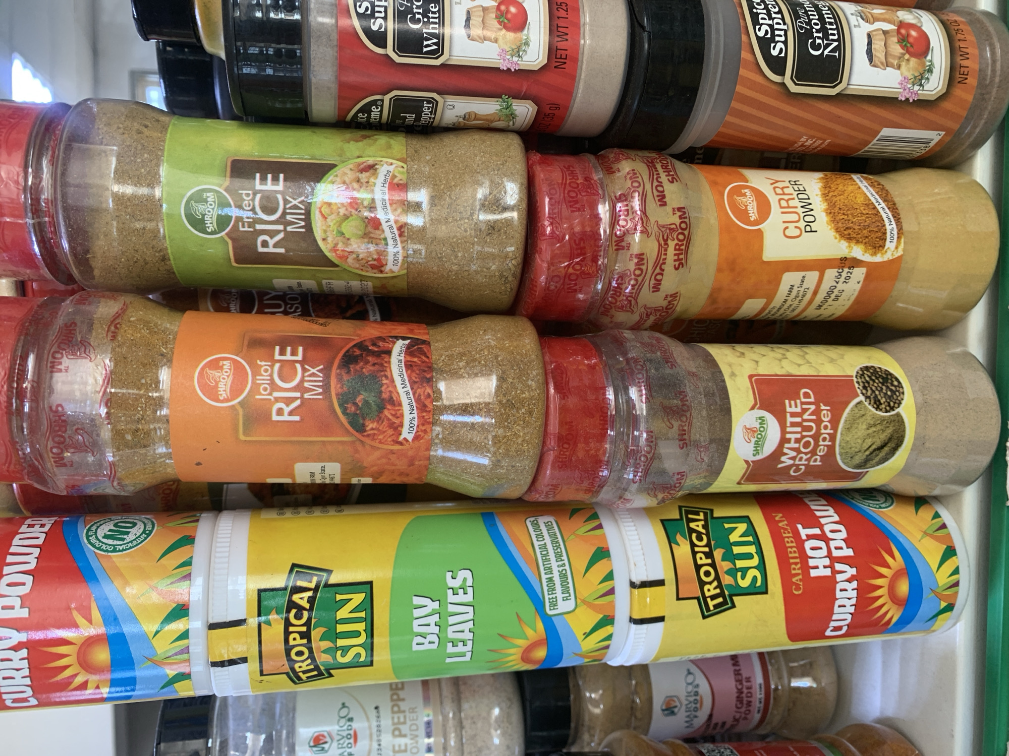 The image displays a shelf filled with various spices in plastic containers. The spices are predominantly in red, yellow, and green packaging. Several brands are visible, including "Tropical Sun," "Jollof Rice Mix," "Fried Rice Mix," "Curry Powder," and "Spice Supreme." The containers vary in size and shape, with some being tall and cylindrical, and others shorter and wider. The shelf itself appears to be a white surface. The lighting suggests an indoor setting. The image is taken from a slightly elevated angle, looking down at the shelf. No people are present in the image.