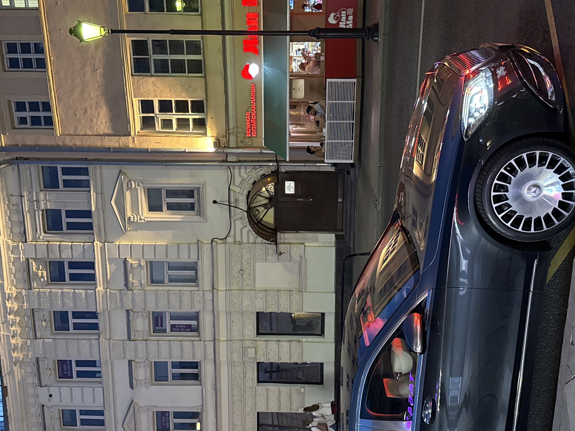 The image shows a street scene in Moscow, Russia, at dusk or night. In the foreground, a dark-colored luxury car, likely a Mercedes-Maybach S-Class based on its distinctive design and wheels, is parked at an angle. The car's headlights are on, and red and purple light reflections are visible on its polished surface, suggesting the presence of other lights or signage.
Behind the car, a two-story building with white and pale yellow facades lines the street. The building features multiple windows, some with ornate detailing. Signs are visible in some windows, indicating "ПРОДАЖА" (Sale) and phone numbers.
To the right of the white building, a separate establishment with a light-colored facade is visible. This building has a bright green awning over its entrance, and a red sign with white lettering above it. The sign appears to read "ВЕЛИКАЯ КИТАЙСКАЯ ЛАПША" (Great Chinese Noodles). Inside this establishment, several people are seated at tables. A street lamp with a green light illuminates the scene.
Further to the right, a red structure with a white logo that resembles a stylized face is partially visible, possibly another part of the restaurant or a neighboring business. The street pavement is visible in the foreground and midground, with a yellow line marking the edge of the road. The overall atmosphere is urban and illuminated by artificial lights.