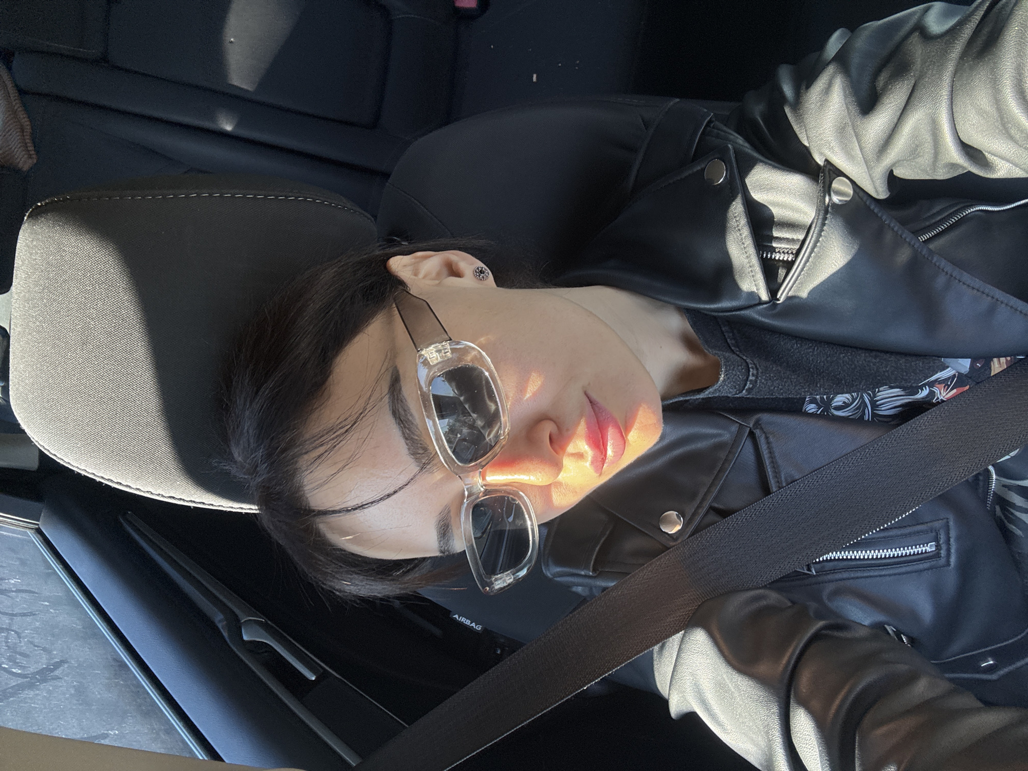 A person is reclining in a car seat, wearing a black leather jacket and clear-framed sunglasses. They are wearing a seatbelt. The interior of the car is primarily black. The location cannot be determined from the given image.