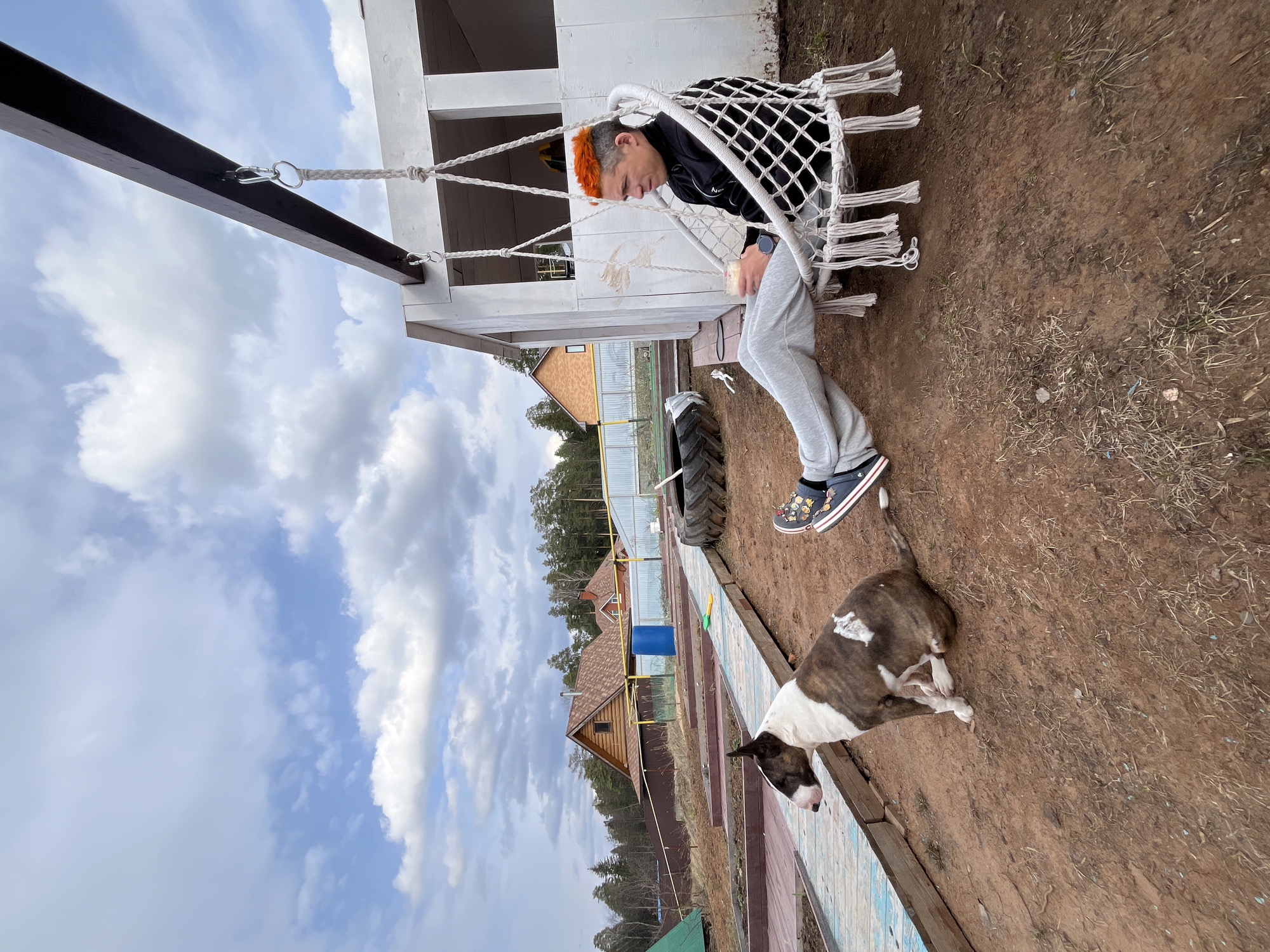 A medium-shot depicts a person sitting in a rope-net swing with a dog lying on the ground. The individual, seen from the side, has bright orange hair and is wearing a black jacket, grey sweatpants, and blue shoes. They are holding a small, possibly disposable, cup. The dog is lying on its side, close to the person.
The swing is attached to a wooden structure. In the background, there are partially constructed buildings and wooden houses, implying an outdoor location, possibly a suburban or rural area. A large tire is also visible nearby.
Based on the location information, the scene is likely situated in or near Chaikovsky, Russia. The environment suggests an outdoor setting.