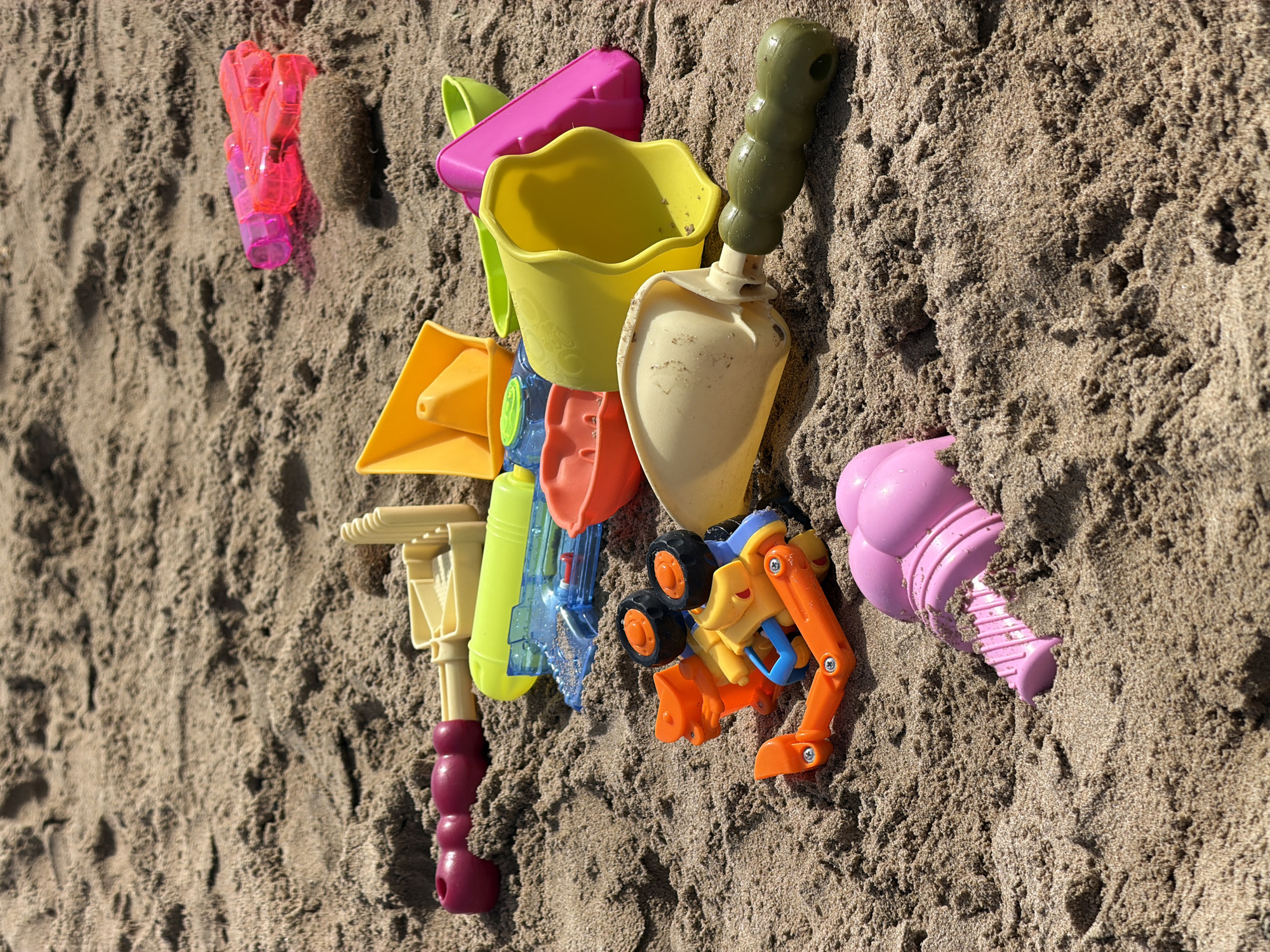 A close-up, high-angle shot shows a collection of colorful plastic beach toys scattered on a sandy surface. The toys, including shovels, buckets, a water gun, and a toy vehicle, are in various shades of pink, yellow, green, blue, and orange. The sand is light brown and appears slightly damp in some areas, with small indentations and textures visible. The background is out of focus, emphasizing the toys in the foreground. The lighting suggests an outdoor setting, likely a beach on a sunny day. There are no people or animals visible in the image. The scene is static and depicts a collection of abandoned or unorganized playthings.