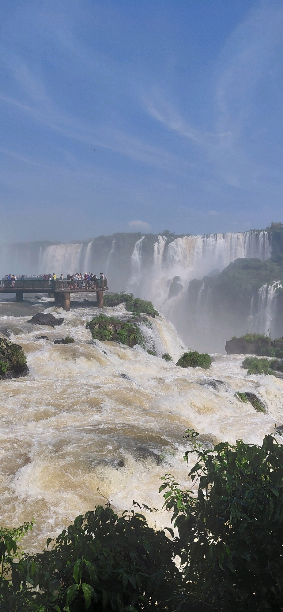 This is a vertical, eye-level, full shot of the Iguazu Falls in Foz do Iguaçu, Brazil. The sky is a bright blue with wispy white clouds. In the foreground, a large area of turbulent, tan-colored water churns. The water is so rough that white foam is visible. Dark green foliage obscures the bottom right corner of the image.
In the middle ground, a raised wooden walkway is visible, spanning left to right. The walkway is filled with numerous people observing the falls. The walkway is supported by sturdy, round pillars that extend into the water. Rocky outcrops covered in green vegetation are scattered throughout the churning water, particularly on the left side of the image.
In the background, a massive, multi-tiered waterfall cascades down a rocky cliff. The water is white and misty, obscuring the details of the cliff face in some areas. The width of the waterfall appears to extend beyond the frame of the image. The overall environment is lush and natural, dominated by water and vegetation.