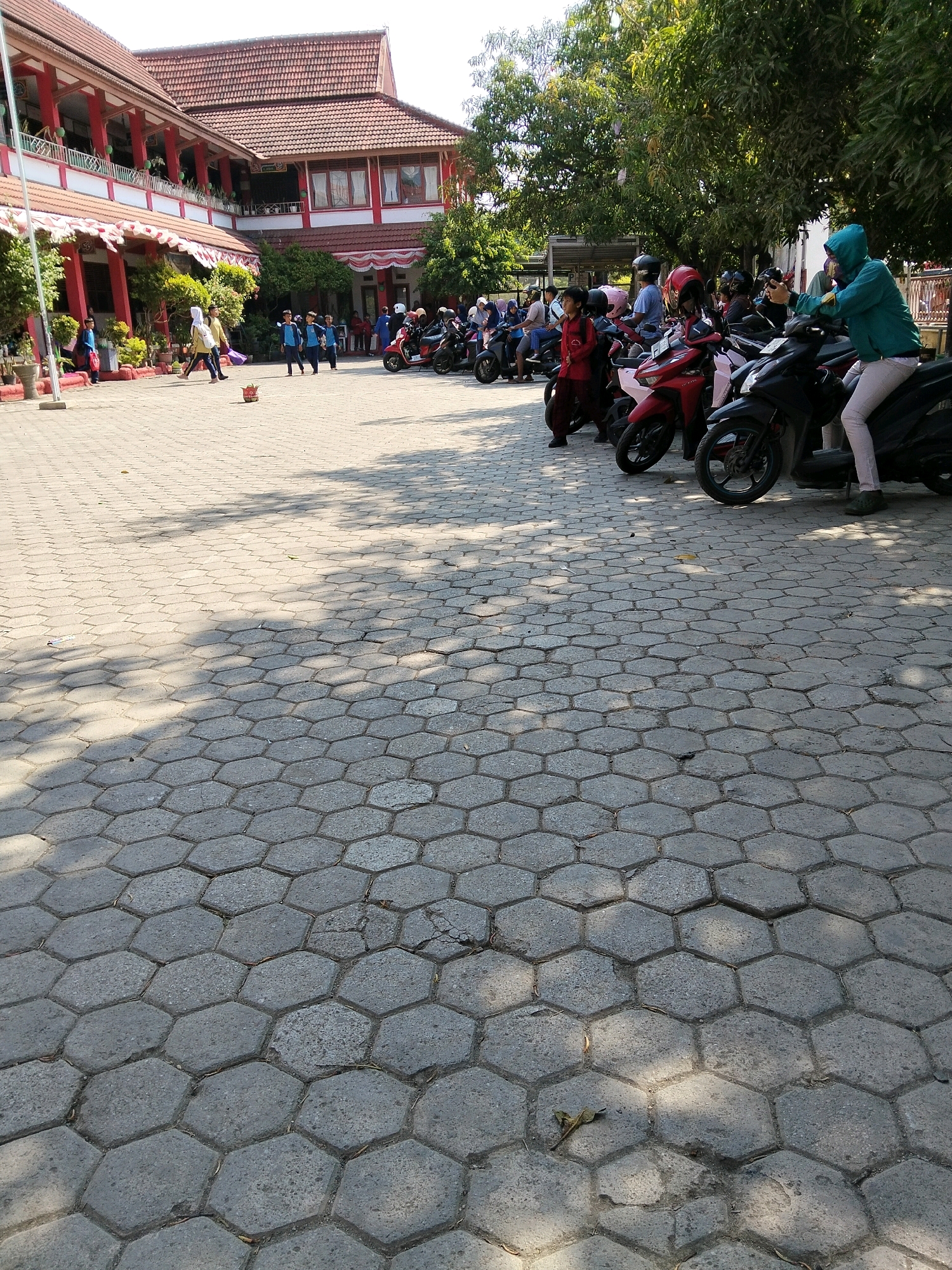The image depicts an outdoor courtyard at a school in Indramayu, Indonesia. The foreground is dominated by hexagonal paving stones, with dappled sunlight and shadows cast by trees overhead. In the mid-ground, a line of parked motorcycles is visible. Several people, including children in blue uniforms, are present. Some are walking, while others are standing near the motorcycles. A two-story school building with red tiled roofs and white balconies forms the background, with lush green trees and foliage surrounding it. A flagpole stands to the left of the building. The scene suggests a typical school day, possibly during a break or after school hours.