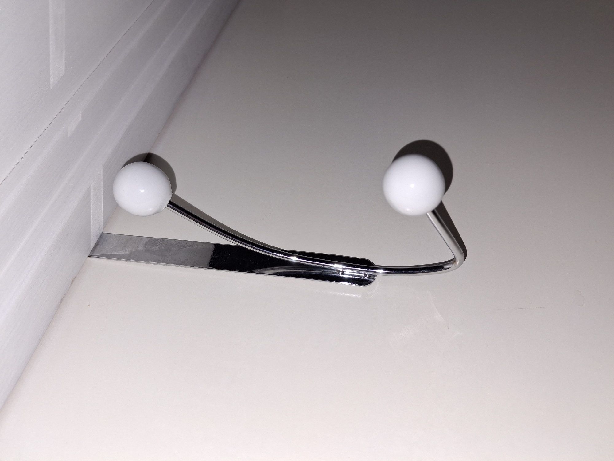 A chrome over-the-door hook with two white ball-shaped ends hangs on a plain, light-colored wall, near a white ceiling. The hook is positioned vertically, with the top hook section resting on the top edge of the door and the curved section extending downwards. The lighting creates shadows on the wall. The image does not depict any people or activities, and the location in Åsgårdstrand, Norway, is not visually represented in the image itself.