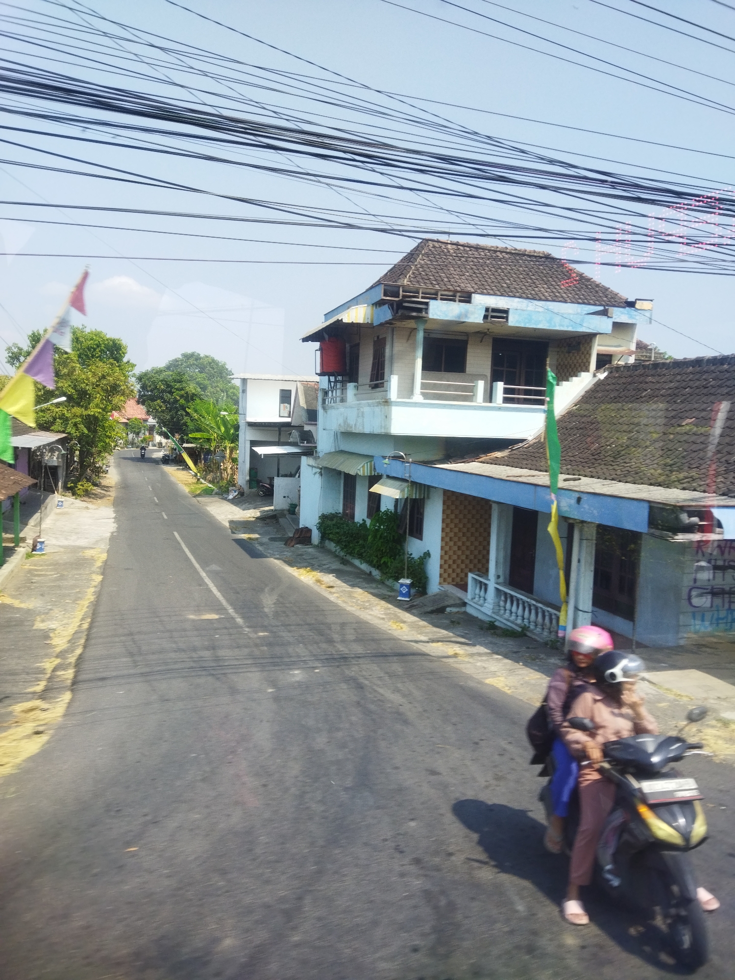 The image captures a street scene in Kepuh, Indonesia, on a clear, sunny day. The road, paved with asphalt and marked with a white line, extends into the distance, flanked by residential buildings and trees. Overhead, a dense network of power lines crisscrosses the sky. In the foreground, two people ride a motorcycle. The person in the back wears a pink helmet and a dark jacket, while the person in the front wears a black helmet and a light brown shirt and pants. They are moving away from the viewer, casting a shadow on the road. Buildings with tiled roofs and blue and white facades line the street, some displaying colorful flags and banners. The environment appears to be a typical semi-urban or rural setting.