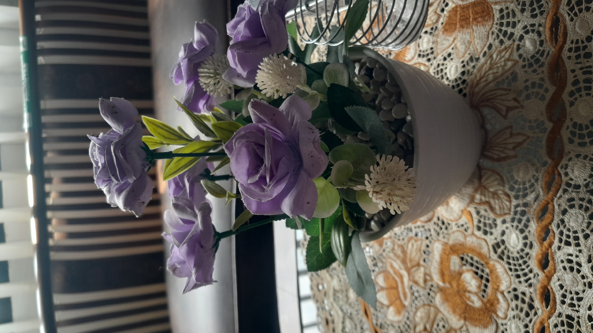 A close-up shot captures a white ceramic pot containing a floral arrangement of artificial purple roses and smaller white flowers. The pot rests on a table covered with a white lace tablecloth adorned with embroidered floral patterns in shades of tan and orange. In the background, vertical blinds are visible, casting striped shadows, suggesting an indoor setting, likely a home or office. The overall scene depicts a decorative display. This scene is located in the city of Tasikmalaya, Indonesia.