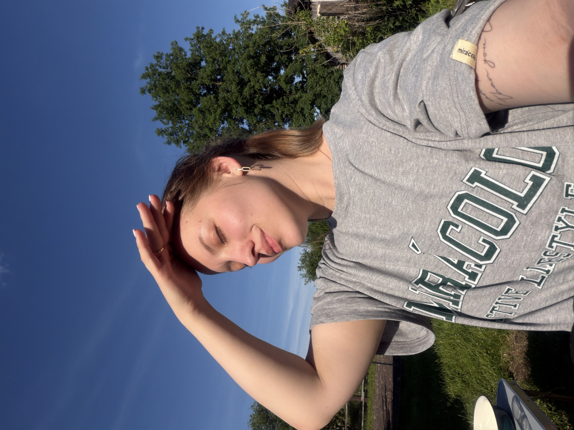 The image captures a light-skinned young adult female outdoors in Reshetnikovo, Russia, under a clear, bright blue sky with sparse, wispy clouds. The female, positioned slightly right of center, has light brown hair pulled back in a low ponytail. She is wearing a grey t-shirt featuring partially visible green and white lettering that reads "MIRACOLL" and "ACTIVE LIFESTYLE". Her left arm is raised, with her hand shielding her eyes from direct sunlight; a thin gold-colored ring is present on her left ring finger, and her fingernails are painted a light color. A gold-colored geometric earring with a dangling element is visible in her right ear, and a faint, light-colored necklace chain is around her neck. On her right forearm, a cursive script tattoo, appearing to end with "than you", is visible. A small, light yellow fabric tag with "miracoll" in lowercase text is attached to the rolled-up right sleeve of her t-shirt. The background consists of vibrant green grass and varied foliage. A large, mature deciduous tree with dense green leaves is prominent in the upper right. To the right of the subject, a rustic wooden structure, possibly a shed or fence, is partially obscured by overgrown vegetation. In the lower left foreground, a small, light-colored outdoor table holds a white bowl, possibly containing a light-colored liquid. The scene is brightly illuminated by direct sunlight, casting visible shadows.