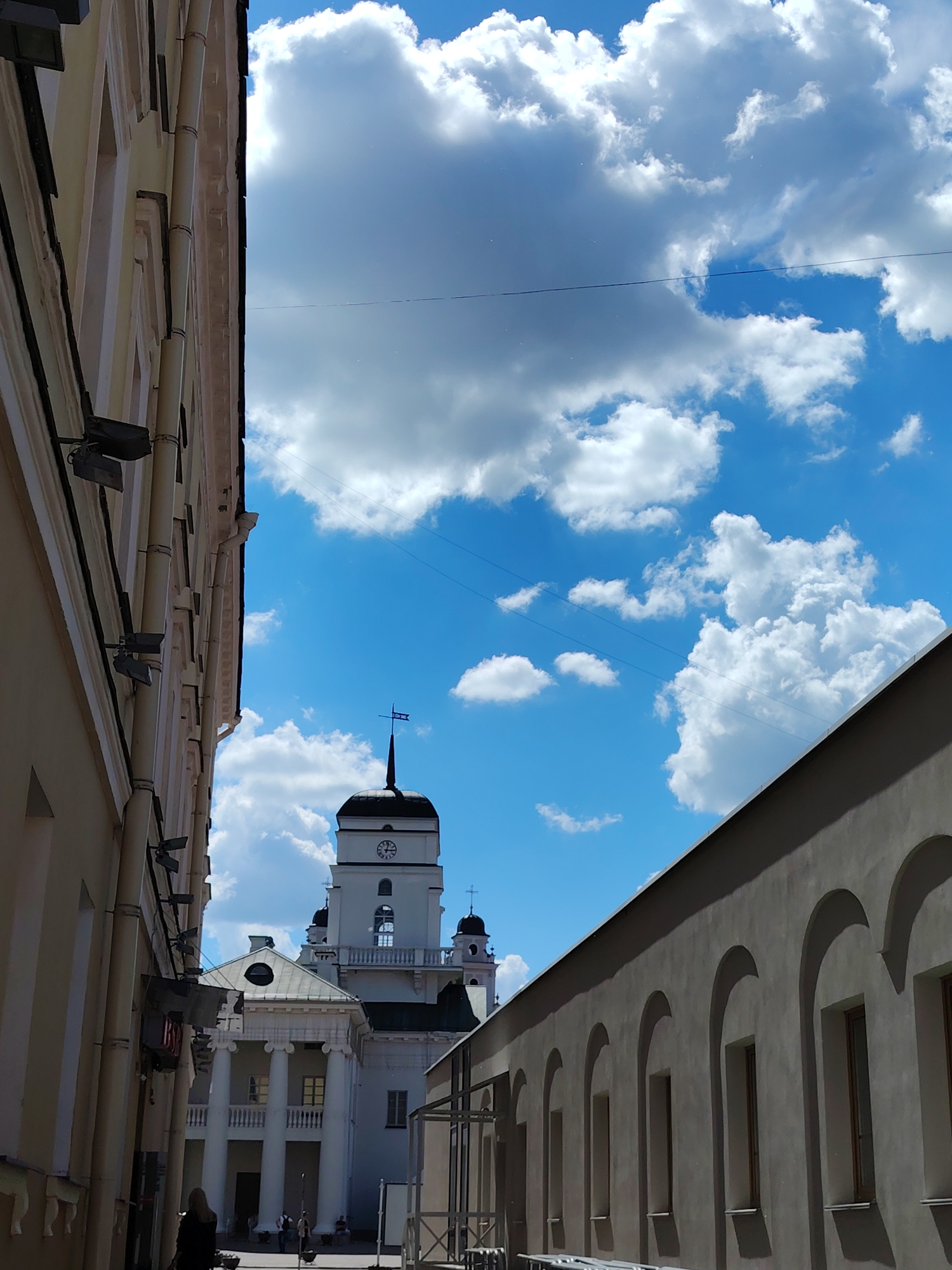 The image presents a vibrant urban scene viewed from a narrow passage, with a prominent historical building under a bright, cloudy sky. On the left, a light-colored building with visible downpipes, windows, and external lighting fixtures runs vertically, creating a canyon-like effect. A red sign with indistinct white text hangs from this building. On the right, a long, light grey building features a series of arched recesses or windows along its length, leading the eye towards the background.
The central focus is a grand white building with classical architecture, featuring a towering clock tower. The tower is topped with a dark, domed roof and a spire that includes a weathervane-like element. The clock face is visible, indicating a time between 10 and 11 o'clock. Below the tower, the building has a substantial portico supported by large columns, suggesting a formal entrance. Further structures with dark domes are visible behind it, implying a larger complex, possibly a church or government building. A few people are seen walking or gathered near the entrance of this central building.
The sky is a brilliant blue, filled with numerous large, fluffy white cumulus clouds, indicating a sunny day. Thin power lines crisscross the open sky between the buildings. The bright daylight and clear weather contribute to a serene and picturesque atmosphere in this historic urban setting, typical of cities like Minsk, Belarus.