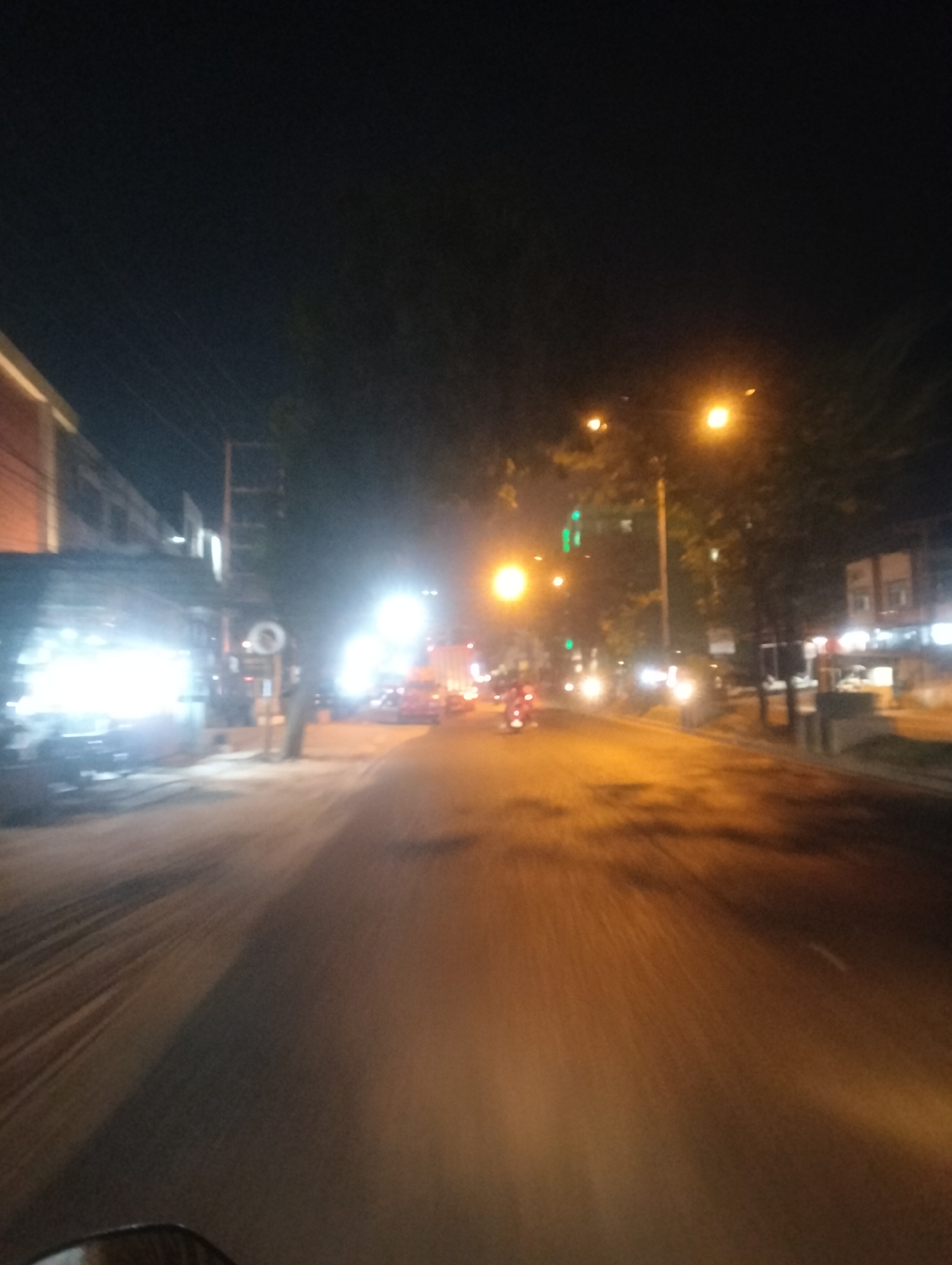 The image depicts a street scene at night, possibly in Air Jamban, Indonesia, characterized by artificial lighting and motion blur. On the left side of the street, there are buildings with illuminated windows and storefronts. A bright, possibly neon, light emanates from one of these establishments. The street itself is paved and appears wet or reflective, with the headlights of vehicles creating streaks of light. A few vehicles, including what appears to be a motorcycle with a rider, are visible on the road. In the background, there are streetlights casting a warm glow and silhouetted trees. The overall impression is of a busy urban environment at night, with the movement captured by the camera resulting in a dynamic and somewhat abstract visual.