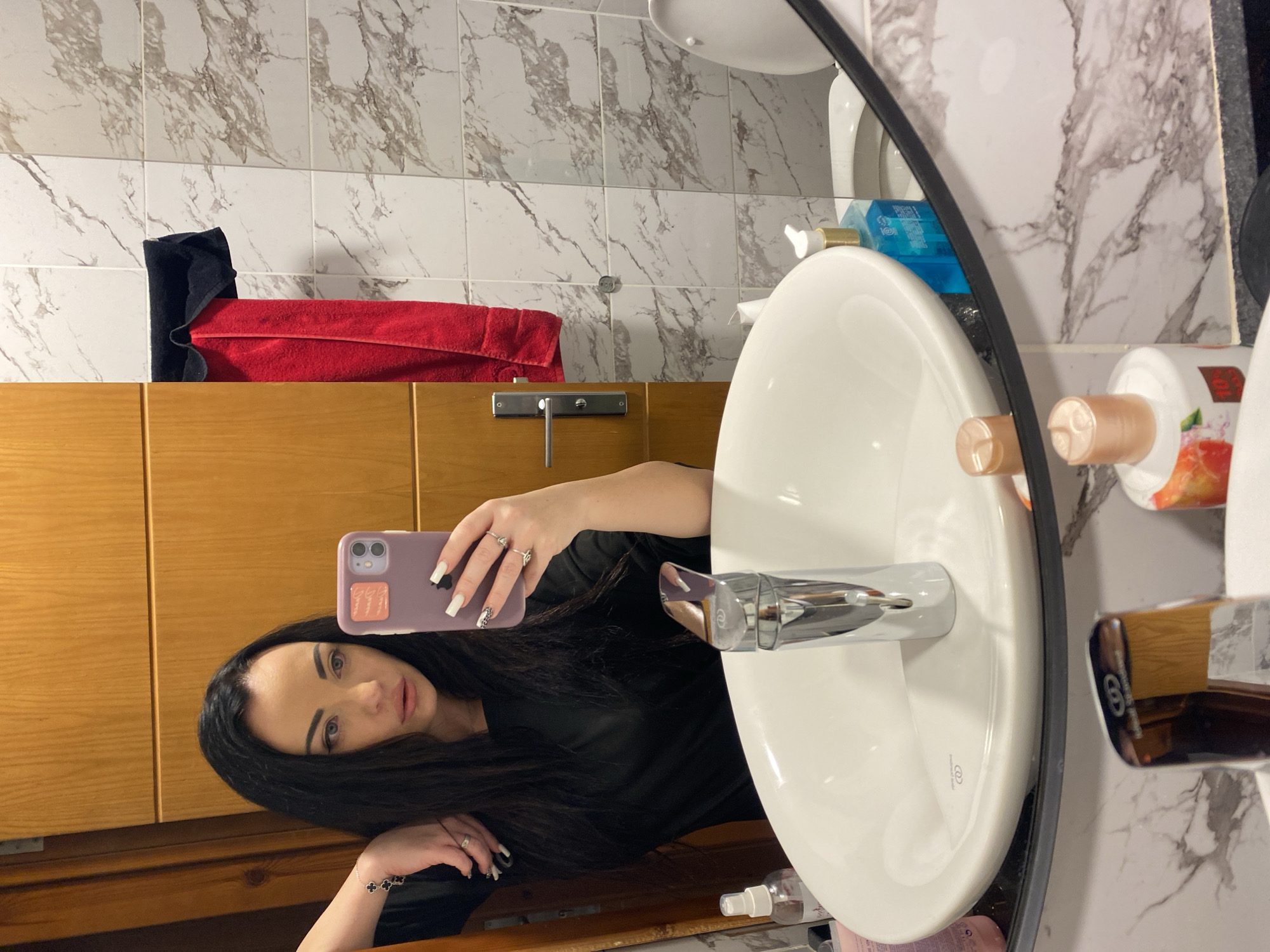 A woman is taking a mirror selfie in a bathroom. She has long, dark hair and is wearing a black shirt. She is holding a purple phone in her right hand, and her left arm is resting on the sink. The bathroom features a white sink, a chrome faucet, and marble-patterned tiles on the walls. A red towel and a black towel are hanging on a rack next to a wooden door. Other items visible in the reflection include various bottles of toiletries, a toilet, and part of a wooden cabinet. The image does not provide enough information to determine the city or country where the photo was taken.