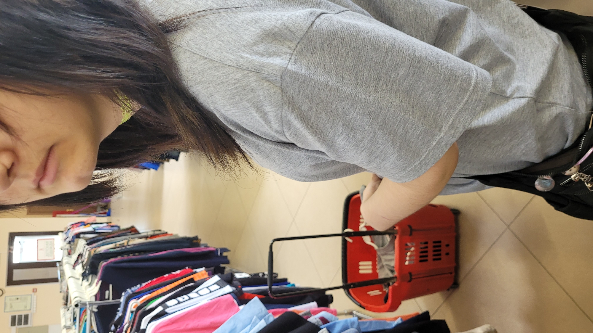 A person in a gray shirt stands in front of a rack of clothes in what appears to be a store in Unknown, Unknown. The person is holding a red shopping basket with black handles. The floor is tiled.