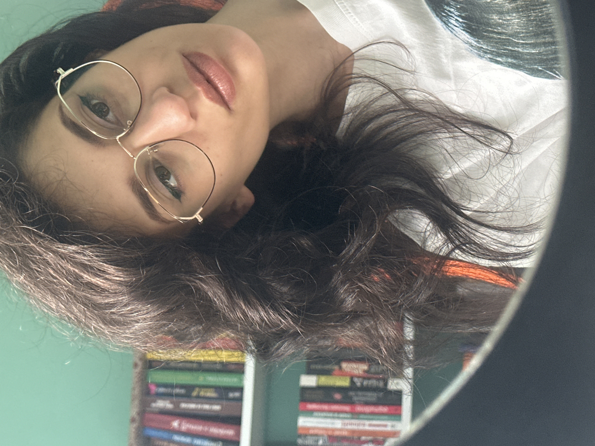 This is a close-up, headshot photograph taken indoors, likely in a home setting given the presence of bookshelves in the background. The subject is a young woman with dark, wavy hair, wearing round, gold-rimmed glasses and a white t-shirt. Her head is tilted, and she is looking directly at the camera. Her lips are parted slightly, and she appears to have makeup on, including eyeliner and lipstick. The lighting suggests it's daytime, with soft light illuminating her face. The background is blurred, but the colors of book spines are visible, indicating a collection of books on shelves. There is no visible text in the image. The overall mood is casual and intimate, perhaps a selfie taken in a personal space. The location context of Sofia, Bulgaria, does not provide any specific visual cues within the image itself.