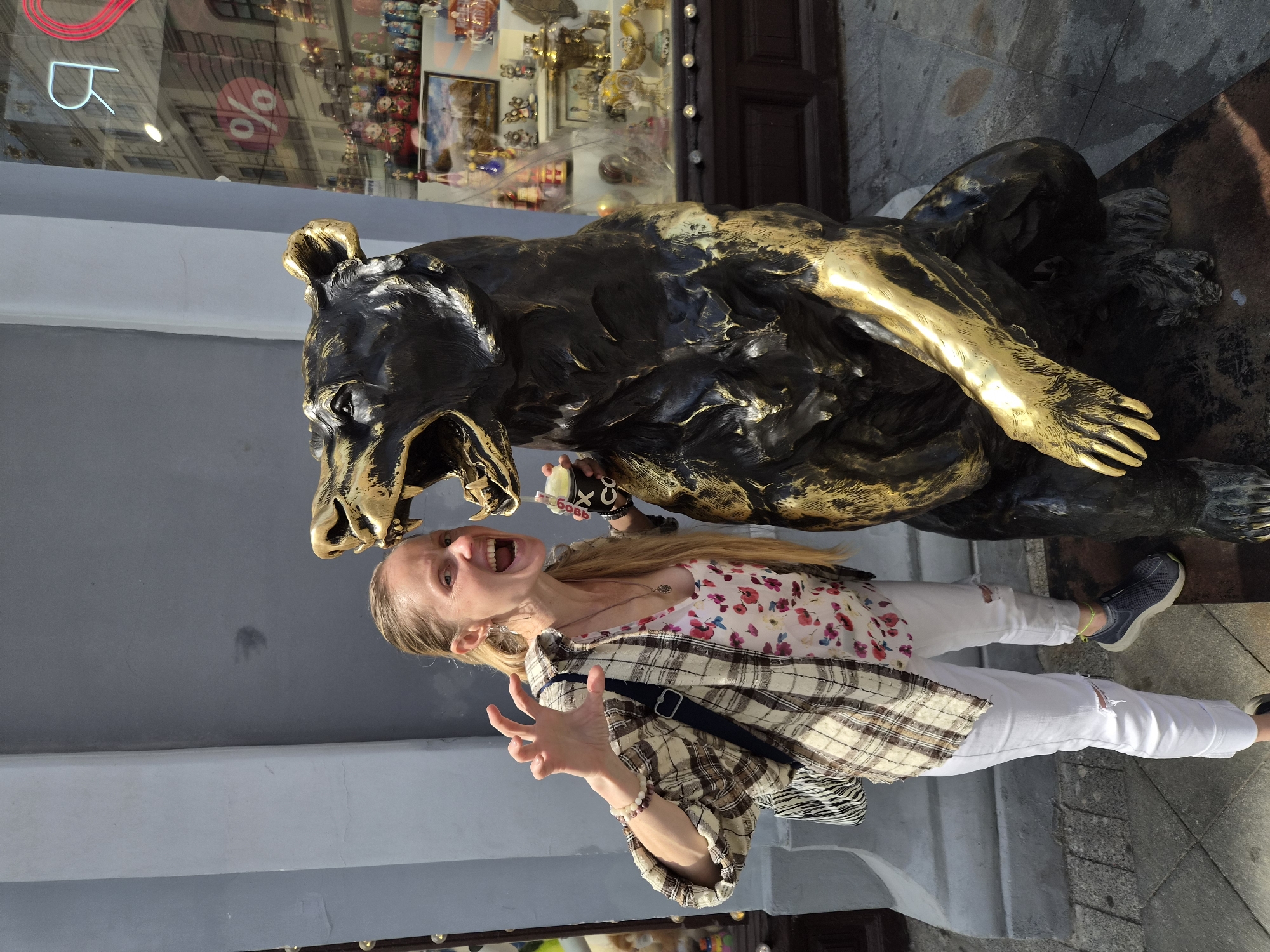 An outdoor, full-body shot depicts a woman interacting with a large bronze bear statue. The woman, with long blonde hair and wearing a white floral tank top, an open plaid button-up shirt, white ripped jeans, and dark sneakers, stands to the left of the statue. She has her mouth wide open in an apparent roar and her left hand raised with fingers splayed, mimicking a claw. In her right hand, she holds a white cup with a drink. The bear statue is rendered in dark bronze with prominent gold detailing on its muzzle, ear tips, chest, and right forelimb, and is posed in an upright, open-mouthed, roaring stance. The background to the immediate left is a plain light gray wall. To the right, a shop window displays a collection of items including Russian nesting dolls, decorative eggs, and samovars, while reflecting a reversed red Cyrillic letter 'Я', a red heart shape, and a white percentage symbol within a red circle. The ground is paved with gray flagstones. The setting is an urban outdoor location in Unknown, Unknown.