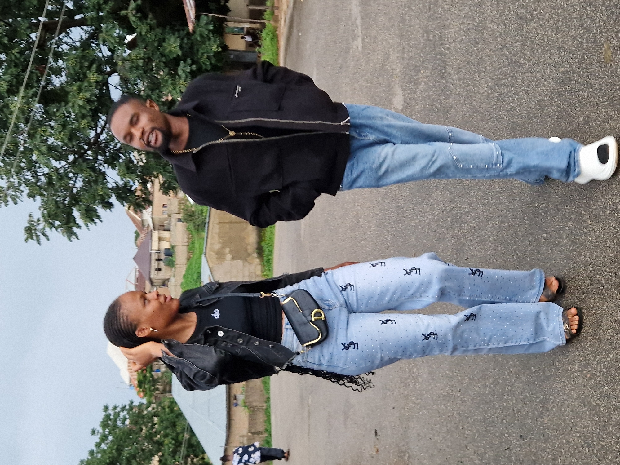Two individuals, a woman and a man, walk side-by-side on a paved road in an outdoor setting in Jos, Nigeria. The woman, on the left, wears a black jacket over a black crop top and light blue jeans adorned with a repeating pattern. She has dark braided hair, earrings, and a black shoulder bag. The man, on the right, is dressed in a black jacket over a black t-shirt and light blue jeans. He is wearing a necklace. Both are walking towards the viewer, and their expressions suggest they are engaged in conversation. The background shows residential buildings, trees, and a cloudy sky, indicative of a typical urban environment.
