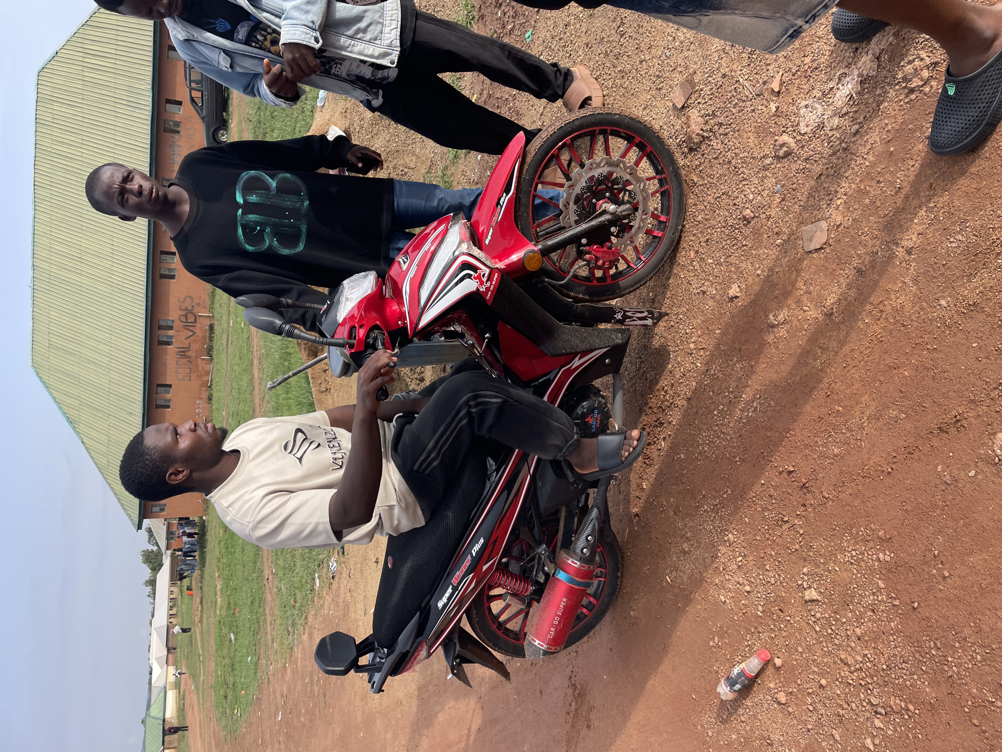 The image shows three young men and a red and black Super Winner Plus motorcycle in what appears to be a semi-urban area in Durimi, Nigeria. One man is sitting on the motorcycle, wearing a light-colored t-shirt and black pants. Another man stands next to the motorcycle, wearing a black sweater with a turquoise design. The third man is partially visible in the top right corner. The ground is a reddish-brown dirt and gravel mix. A plastic bottle lies on the ground nearby. A building with a light green roof is visible in the background. Other buildings and greenery are also visible in the distance. The motorcycle is parked, and the man on it appears to be stationary. The other two men are standing and appear to be interacting with each other and the motorcycle.