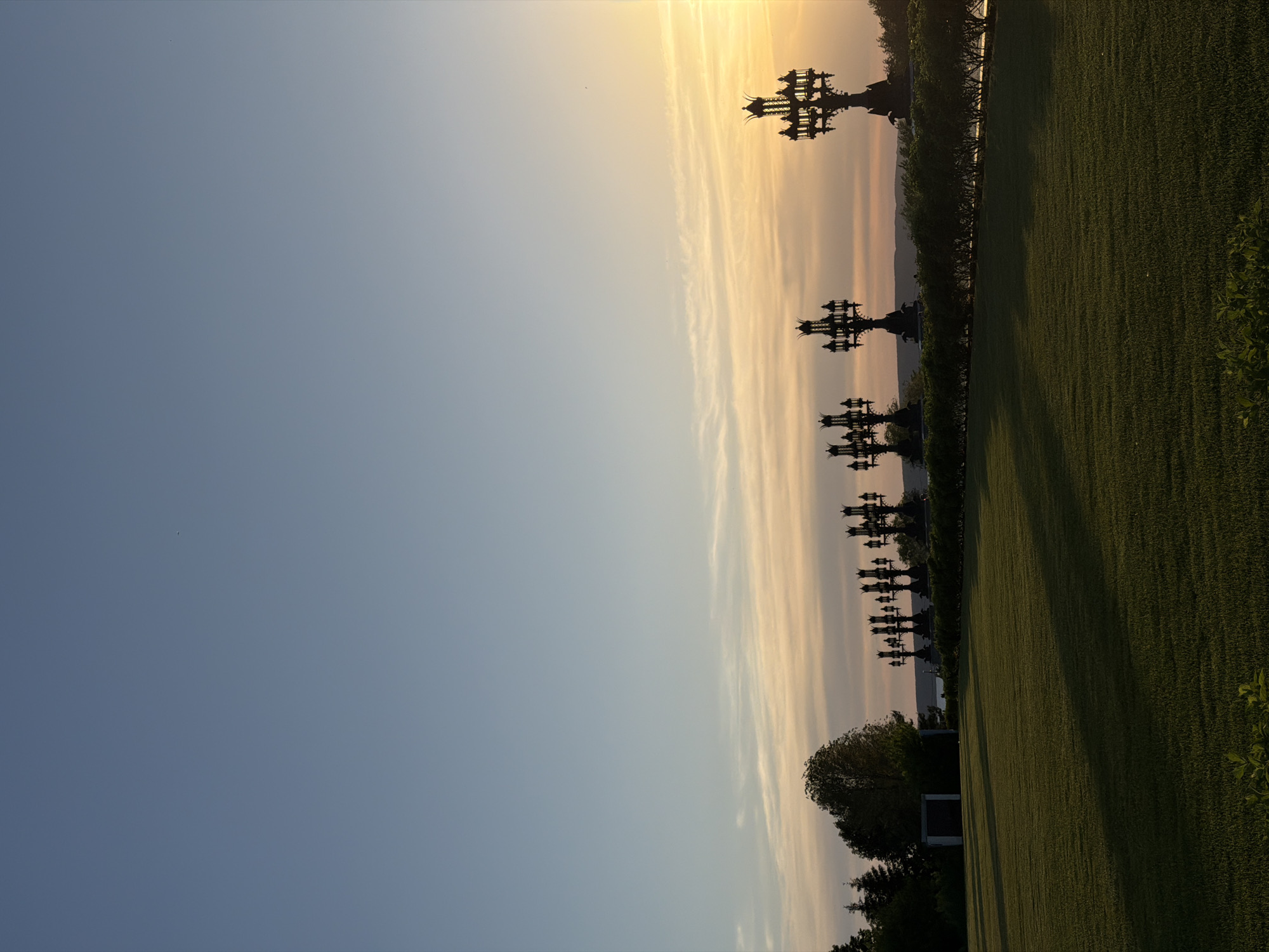The image depicts a row of elaborate metal crosses silhouetted against a sunset sky, with a grassy field in the foreground. The location is Khryashchevka, Russia. The crosses are positioned along the edge of the field, behind a hedge. The sky transitions from blue to orange and yellow near the horizon. Some trees can be seen near the crosses and the field.