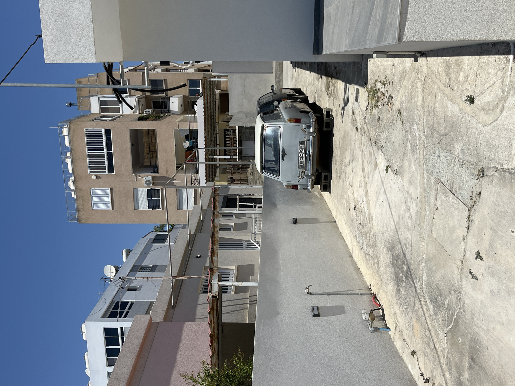 This is a tilted, full-shot, eye-level media file of an urban scene with residential buildings and parked cars under a clear blue sky. The scene appears to be in a courtyard or alleyway between buildings. In the foreground, a light-colored wall occupies the right side, casting a shadow. A concrete ground is visible, with cracks and some sparse vegetation growing from it. Two cars are parked on the concrete: a light-colored vintage station wagon with a license plate reading "CE929" and a darker car behind it.
The background features several multi-story apartment buildings with beige and grey facades, balconies, and numerous windows. Some windows have white shutters. Air conditioning units are visible on some of the buildings. Antennas and satellite dishes are also mounted on the rooftops and walls of the buildings. The overall setting is a dense urban environment, likely a residential area. The lighting suggests it is daytime, and the clear sky indicates fair weather. The image conveys a sense of everyday life in a Mediterranean city, with no specific activity or event taking place beyond the cars being parked. The scene is static and focuses on the architecture and urban landscape.