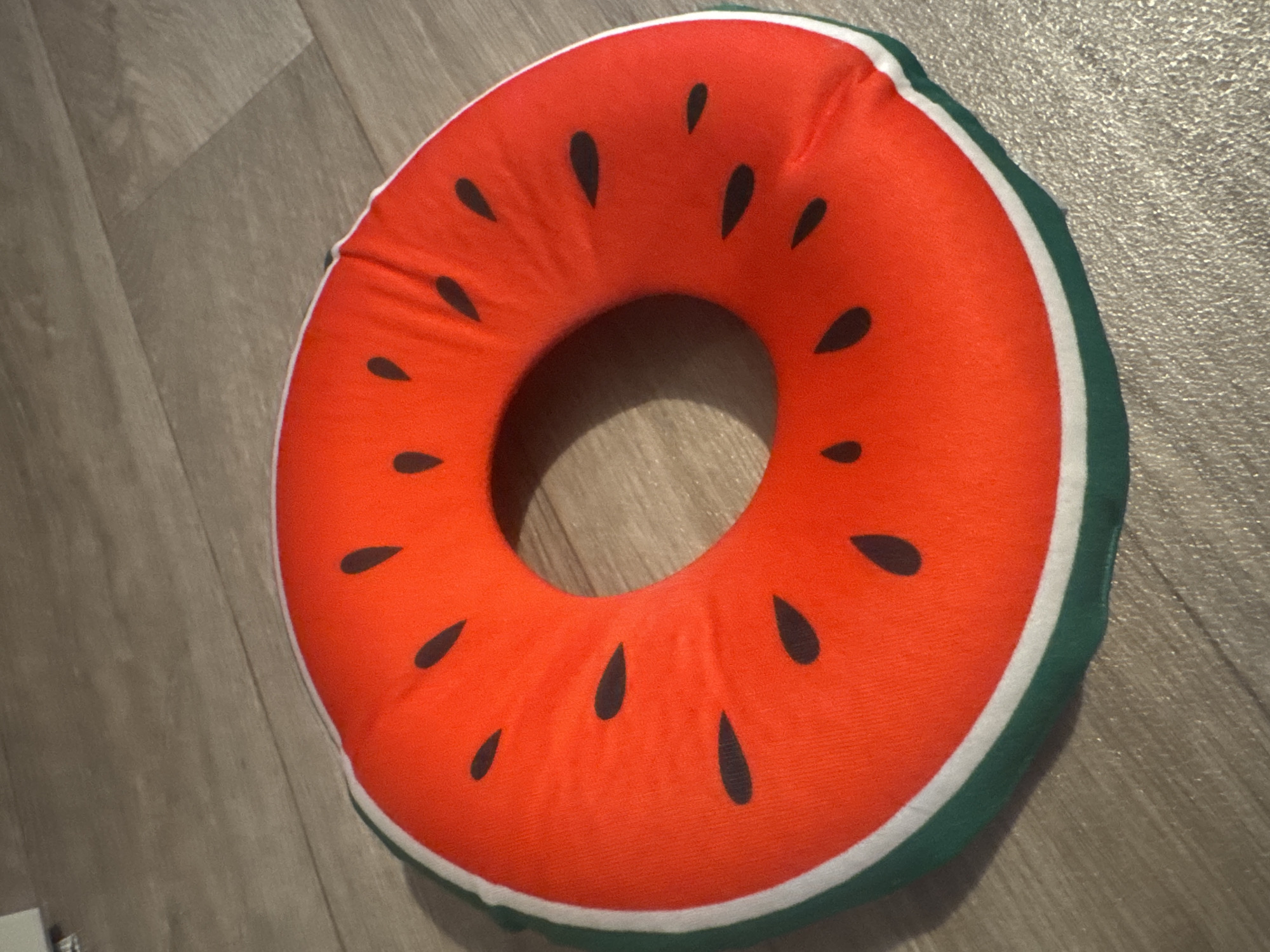 An overhead shot shows a round, inflatable object resembling a slice of watermelon, resting on a wooden floor. The object has a bright orange interior with scattered dark, teardrop-shaped "seeds," and a green outer ring with a white stripe between the orange and green. The floorboards are oriented diagonally from the top left to the bottom right of the frame.