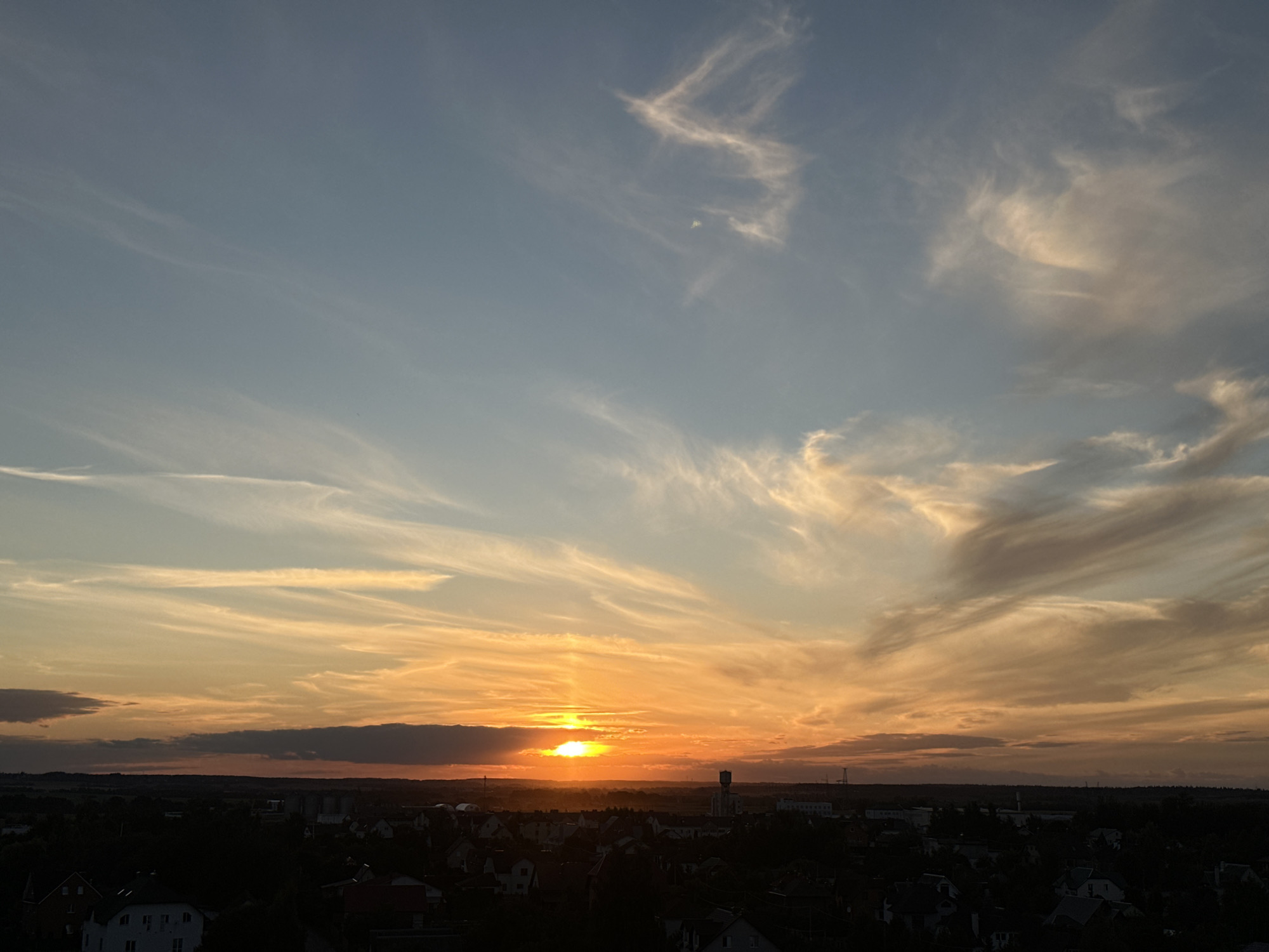 This image depicts a sunset over a town, likely located within the territory of Шчомысліцкі сельскі Савет, Belarus. The sky is a gradient of colors, with pale blue at the top transitioning to vibrant orange and yellow near the horizon. Wispy clouds streak across the sky, catching the warm light of the setting sun. The sun itself is partially obscured by a dark band of clouds low on the horizon, but its rays are still visible, creating a luminous effect. Below the sky, a silhouetted landscape of a town is visible. The dark shapes of buildings, trees, and some industrial structures are discernible. A prominent water tower is visible in the mid-ground of the town. The overall impression is one of a tranquil evening scene. There are no people or animals visible in the image.