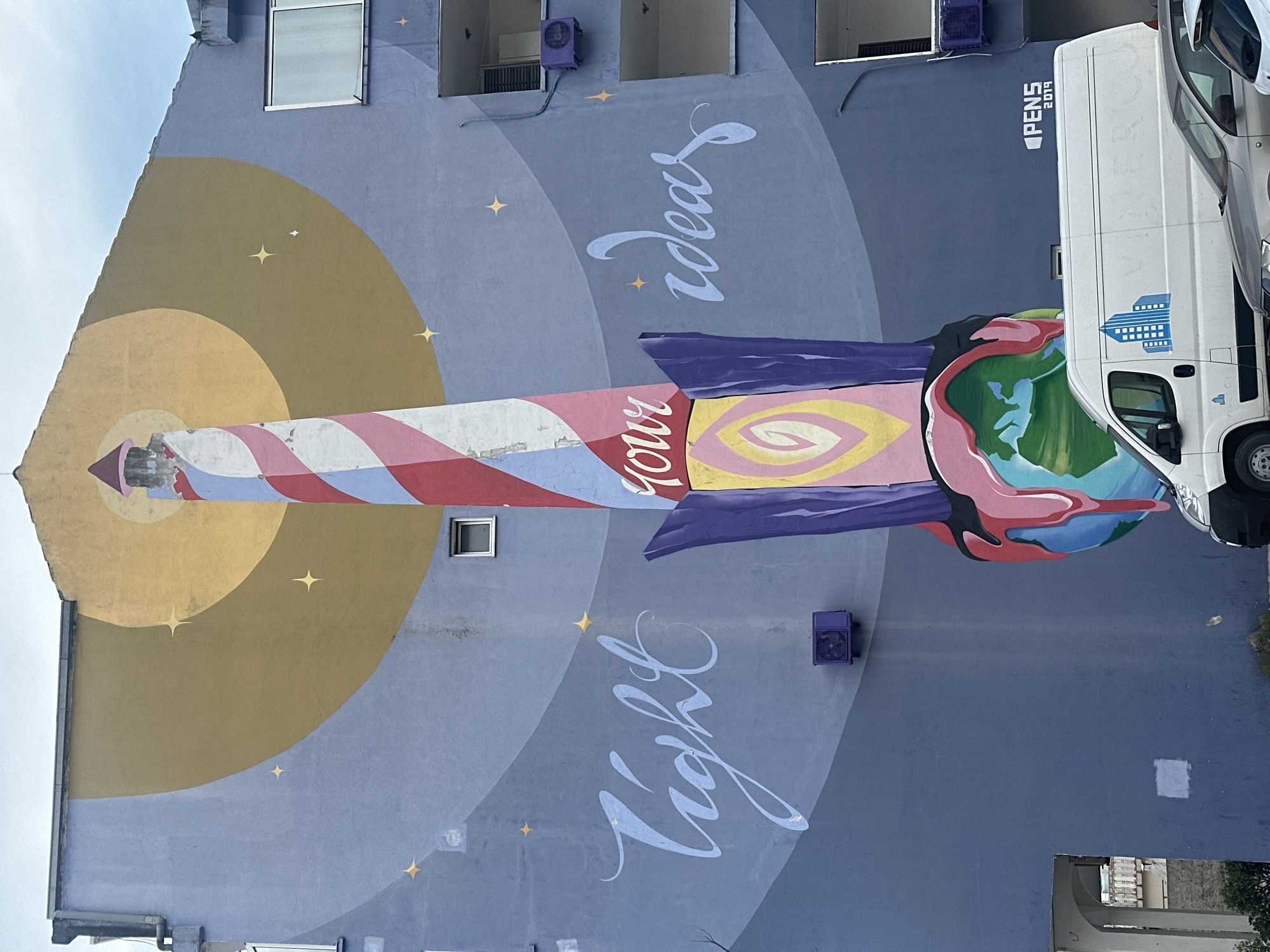 A large mural adorns the side of a building in Budva, Montenegro. The mural features a stylized lighthouse emitting golden light, with text reading "Light your ideas." A colorful, rocket-like structure emerges from the lighthouse, ending in an abstract representation of the earth. The building is a multi-story structure with visible windows and air conditioning units. A white van with "PENS 2014" written on it is parked in front of the building, partially obscuring the mural.