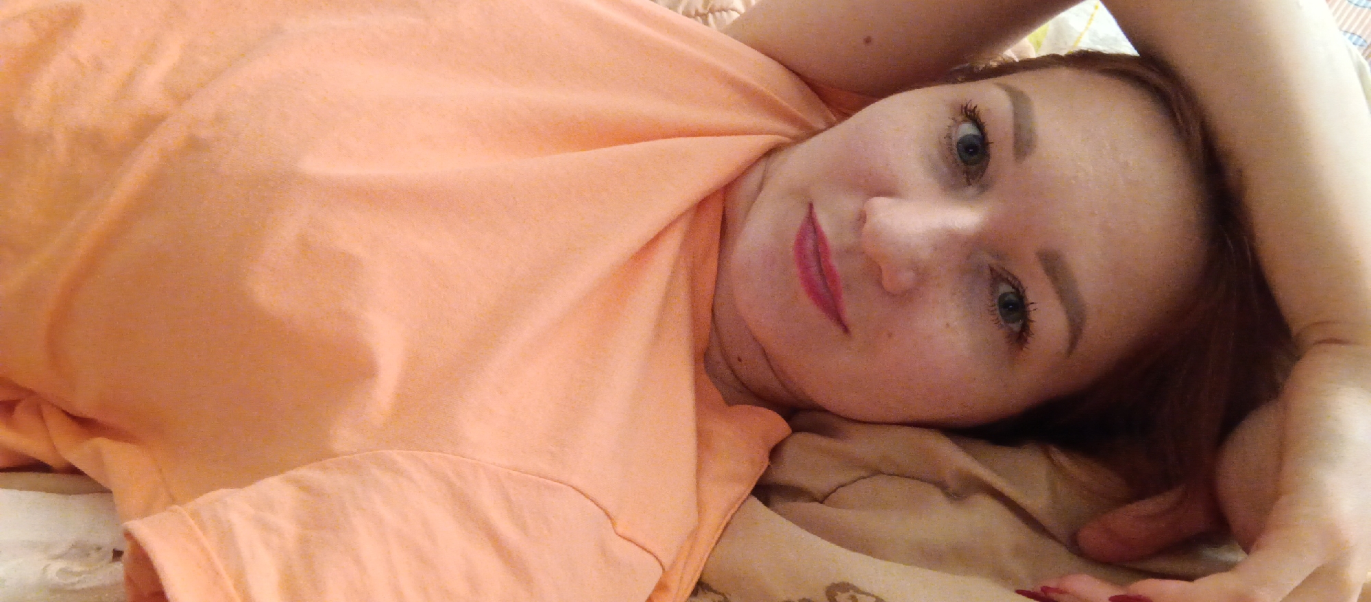 An image captures a woman lying down, looking directly at the camera. She appears to be indoors, possibly on a bed or sofa, indicated by the soft, patterned fabric beneath her head and left arm. Her head is tilted, and she is positioned diagonally across the frame.
The woman has light skin with visible freckles and blue-green eyes. She has reddish-brown hair, some of which is visible around her head. Her makeup includes dark eyeliner or mascara on her eyelashes, well-defined eyebrows, and bright pink lipstick. Her expression is neutral and direct.
She is wearing a simple, light-colored (peach or light orange) t-shirt or casual top, which drapes loosely around her upper body. Her right arm is raised and extended above her head, while her left arm is partially visible beneath her. Her fingers are visible at the bottom right, showing red nail polish. The background is mostly blurred, showing more of the soft, light orange fabric of her clothing and a darker, patterned fabric of what appears to be a blanket or cushion. There is no visible text in the image.