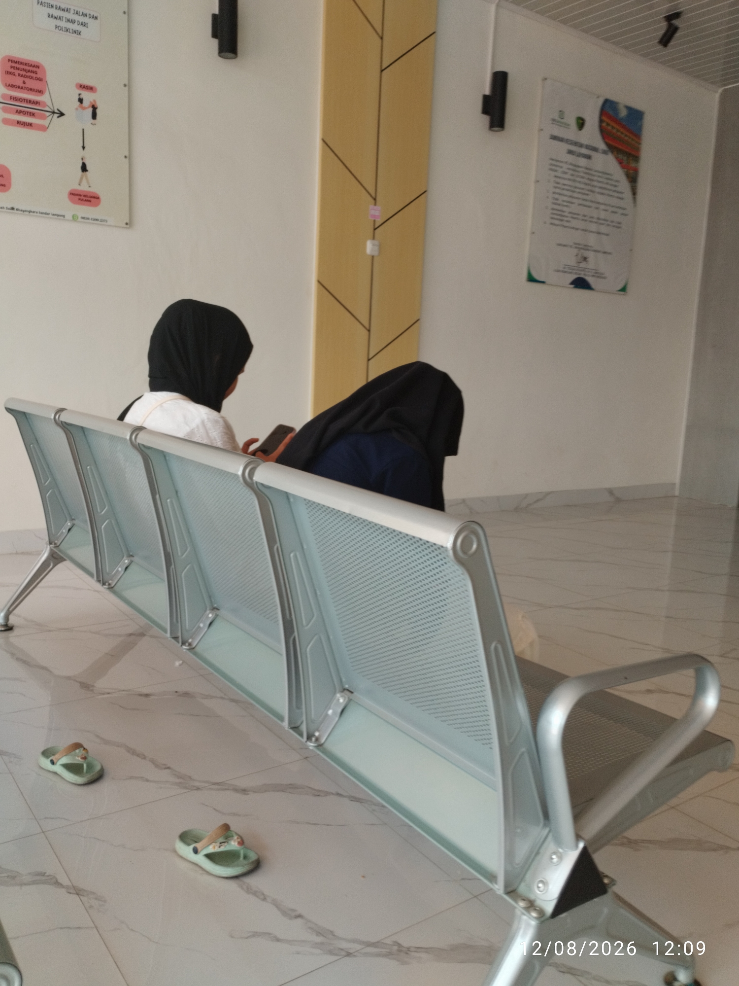 In an indoor waiting area, possibly in the city of Bandar Lampung, Indonesia, two individuals wearing headscarves are seated on a light-colored, metal bench. The person on the left is wearing a white top and a black headscarf, and is looking at their phone. The person on the right is wearing a dark-colored headscarf and a navy blue top, and is also looking down. The floor is tiled with a light marble pattern. Two pairs of child-sized, light green flip-flops are on the floor in front of the bench. The walls are painted white, with a section of yellow wooden paneling behind the individuals. A sign with a flowchart is visible on the wall to the left, and another poster is mounted on the wall to the right. A date stamp reads "12/08/2026 12:09".