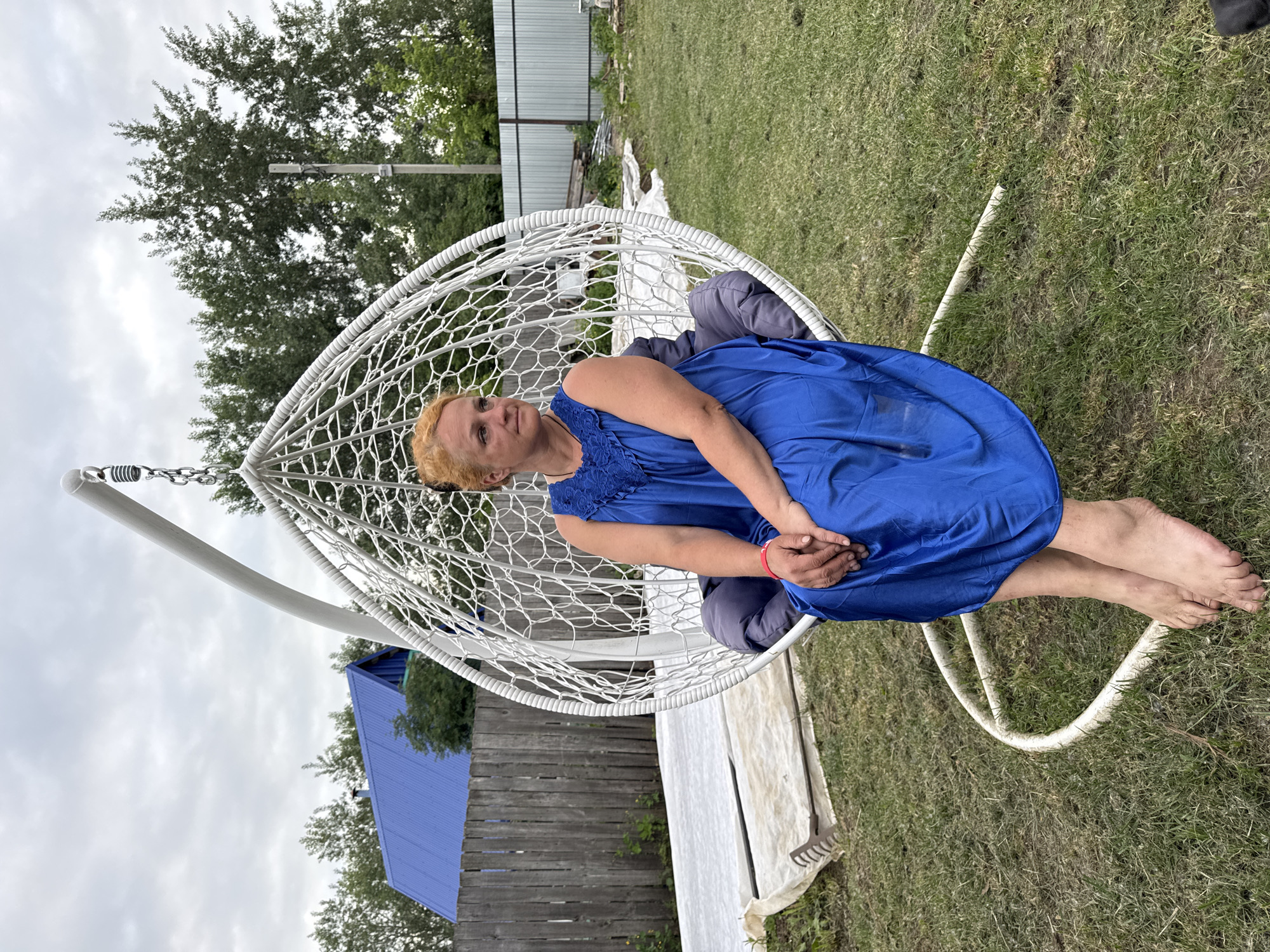 An adult woman with short, red hair and fair skin is sitting in a white, egg-shaped hanging chair outdoors. She is wearing a long, royal blue dress with lace detailing around the neckline. The woman is barefoot, and her hands are clasped in her lap. She is looking to her right. The hanging chair is supported by a white metal stand. The background shows a grassy yard with trees, a wooden fence, and a building with a blue roof. The sky is overcast. This scene appears to be in a residential area, possibly a dacha or country house, in Chaikovsky, Russia.
