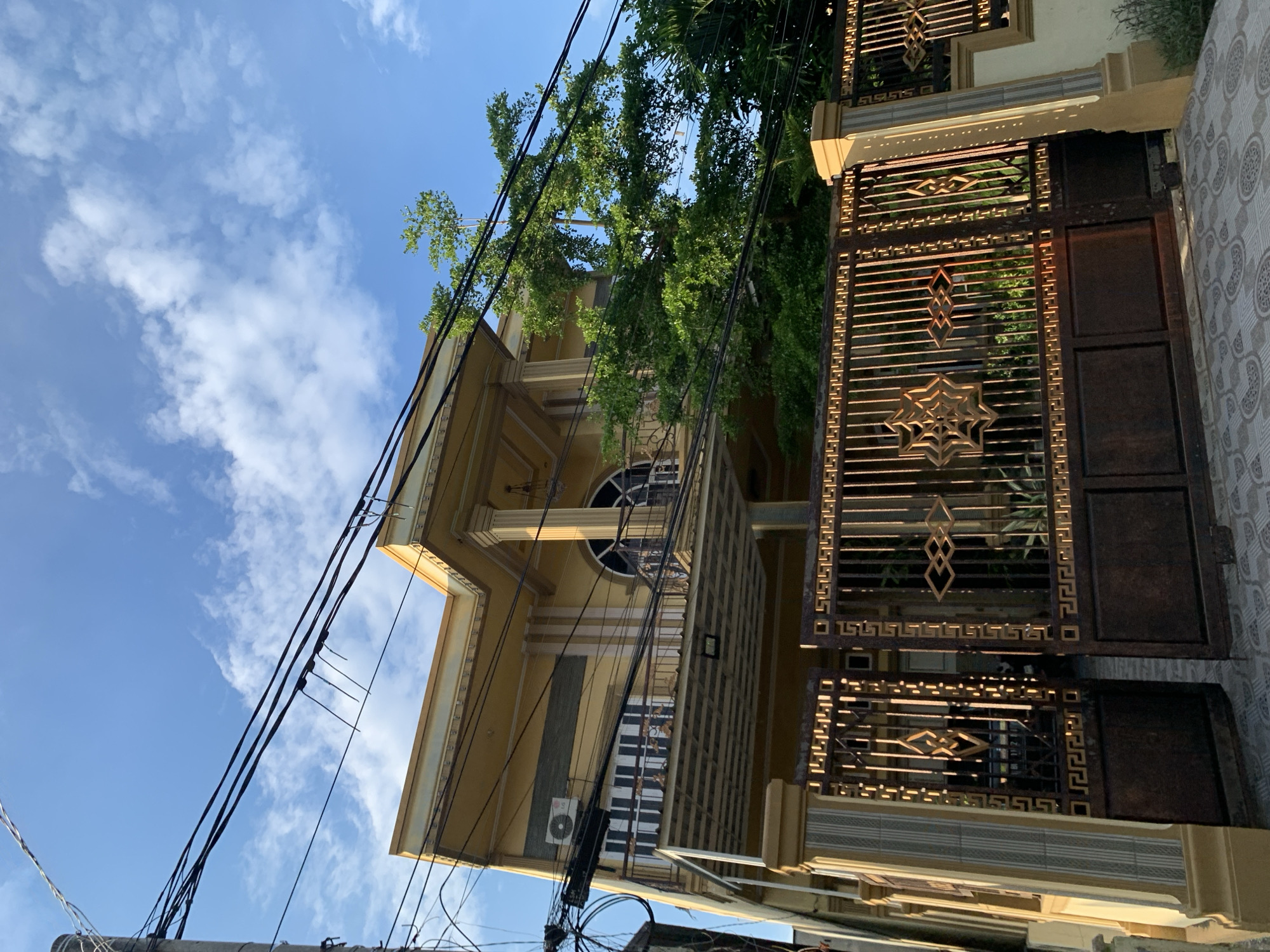 The image captures the facade of a two-story yellow house located in the city of Lhokseumawe, Indonesia. The house features a prominent balcony with a metal railing on the second floor, accessible through large doors. The ground floor has a covered entrance and a decorative metal gate that is partially open, revealing a glimpse of the property inside, including a parked vehicle. Overhead, a tangle of electrical wires crosses the frame, suggesting the presence of a utility pole. Lush green trees partially obscure the side of the house and the fence. The sky above is blue with scattered white clouds. The pavement in front of the gate displays a patterned tile design. The overall scene depicts a residential property in what appears to be a tropical climate.