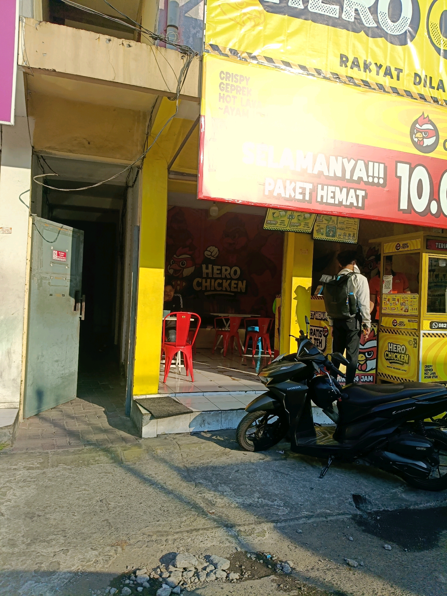 In Surabaya, Indonesia, a street-level view captures the storefront of "Hero Chicken." A vibrant yellow awning stretches across the facade, emblazoned with "RAKYAT DILAYANI" (The People are Served). Below this, a red banner proclaims "SELAMANYA!!!" (Forever!!!) and "PAKET HEMAT" (Economical Package), with a prominent display of "10.0" suggesting a price. Inside the store, red chairs are visible, and a person is seated at a table. To the right, a yellow food stall, also branded "Hero Chicken," has a counter where a person in an orange uniform serves customers. A man wearing a white shirt, dark pants, and a backpack stands in front of the stall, facing away from the camera. A black Honda motorcycle is parked in the foreground. The environment appears to be a commercial street, with buildings and a paved walkway. The sun casts shadows, indicating it is daytime.