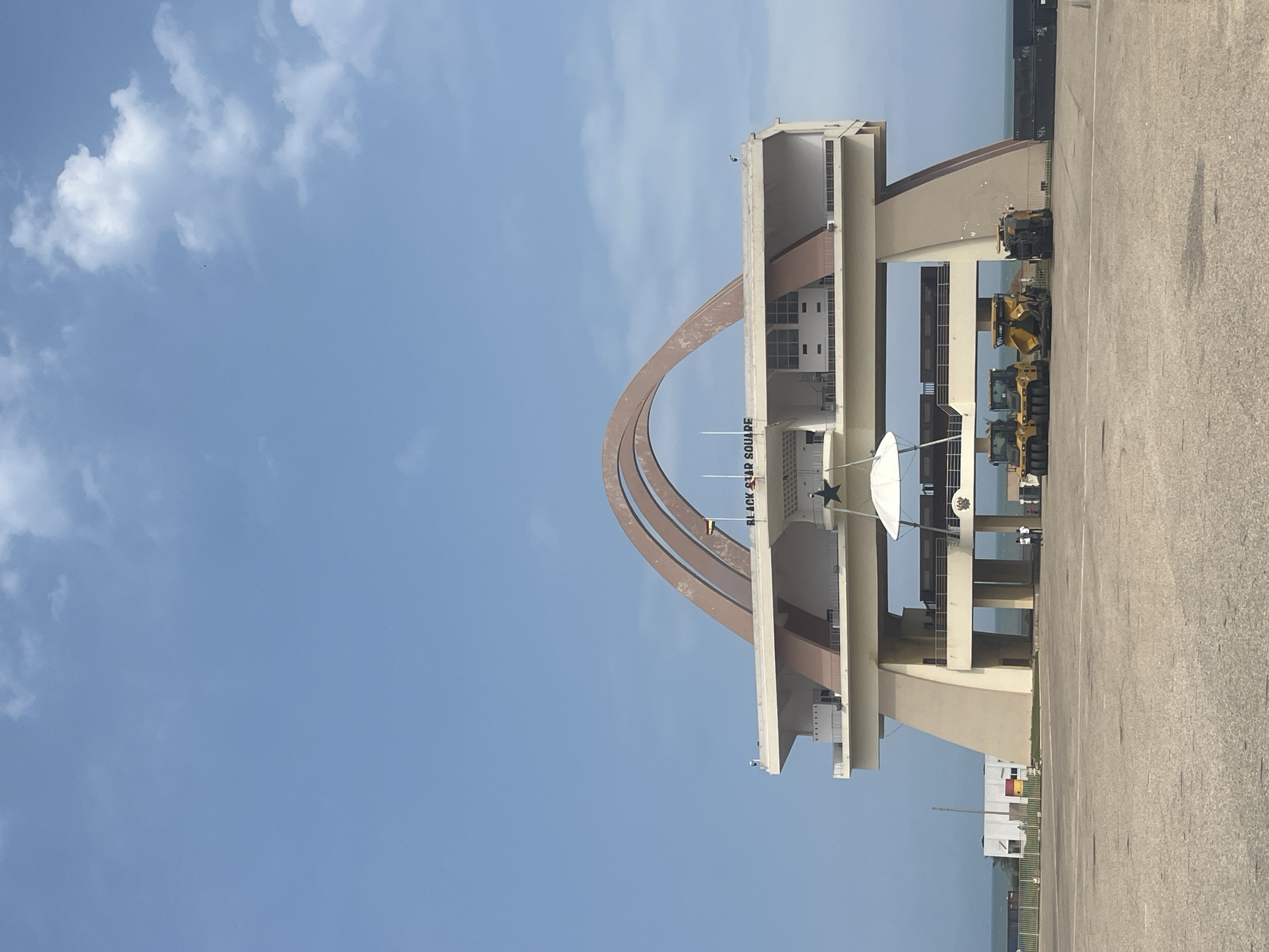 This image depicts the Independence Arch at Black Star Square in Accra, Ghana. The arch, a prominent landmark, is a monumental structure with large, curved beams supporting a platform. The words "BLACK STAR SQUARE" are visible on the platform. Below the platform, there are several levels with windows and railings. A Ghanaian flag flies from a flagpole on the right side of the structure.
In the foreground, a large, paved open area is visible, likely a public square. Several pieces of construction equipment, including yellow rollers and an asphalt paver, are parked on the pavement. A few people are standing near the construction equipment and under the arch. The ocean is visible in the background, with a faint horizon line. The sky is clear blue with scattered white clouds.