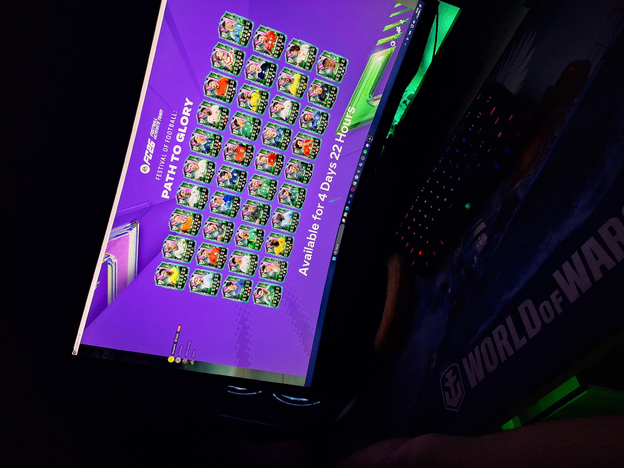 A dimly lit indoor scene captures a desktop computer setup, presumably located in the city of Varna, Bulgaria, though the exact specific location within the city is not discernible from the image. The central focus is a computer monitor displaying the interface of the video game 'EA Sports FC26,' specifically the 'FESTIVAL OF FOOTBALL: PATH TO GLORY' event within Ultimate Team. The screen presents a grid of numerous virtual player cards, predominantly featuring male football players with ratings ranging from 91 to 96, including identifiable players like Vinicius Jr., Frenkie De Jong, Rodrygo, Antonio Rudiger, and Luka Modric. A digital timer at the bottom of the screen indicates the event is "Available for 4 Days 22 Hours." The Windows 11 taskbar is visible at the bottom right, showing icons for search, a web browser, Spotify, Discord, and system indicators including the time (23:55) and date (7.6.20XX). On the upper left side of the screen, a partial chat window or friend list displays usernames such as 'Spongebob' (with 'TCAP' and 'LIVE' indicators), 'BrianCheng,' 'miszka_amusanki,' and 'Stykala.'
To the immediate left of the monitor, a dark-colored computer tower features prominent vertical cool blue/cyan LED lighting strips. In the foreground, a dark mechanical gaming keyboard with multi-color RGB backlighting is positioned on a large, dark mousepad. The mousepad displays the text "WORLD of WAR" alongside an emblem, likely indicating "World of Warships." A green light source is visible beneath the desk on the left side of the frame. A person's bare forearm and hand are partially visible in the lower-left corner, resting on the desk or an armrest.