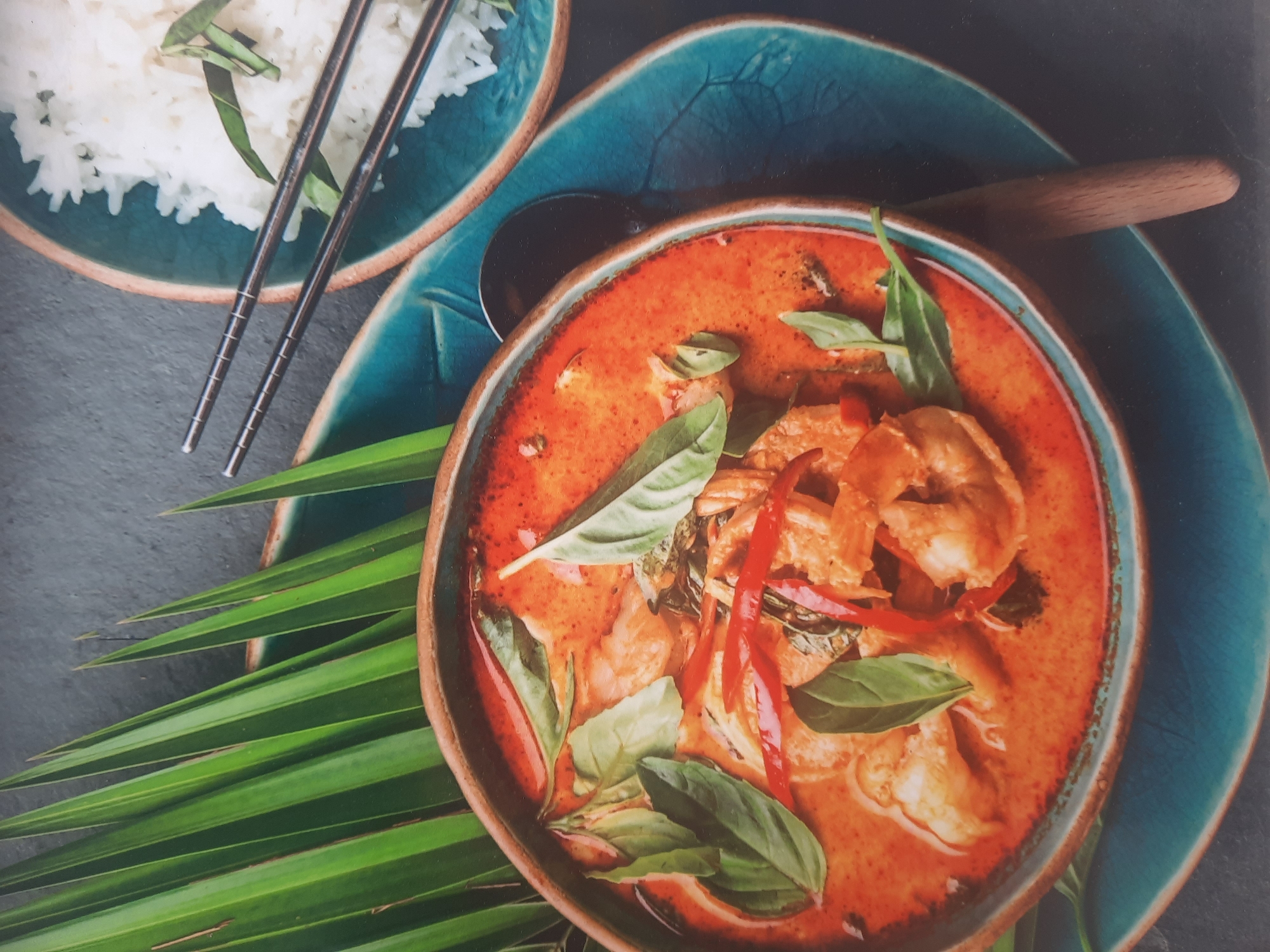 This is a close-up, overhead shot of a bowl of Thai red curry with shrimp, served with a side of white rice and chopsticks. The curry is a vibrant orange-red color and is garnished with fresh basil leaves and sliced red chili peppers. The rice is fluffy and white, and the chopsticks are made of silver metal. The meal is served on a dark blue ceramic plate with a leaf pattern. There are also large green palm leaves in the background, adding a tropical feel to the scene. The setting appears to be a restaurant or home, but there is no specific information to pinpoint the location in Rosenheim, Germany. The lighting is natural and soft, creating a warm and inviting atmosphere. The focus is sharp on the food, highlighting its texture and colors.