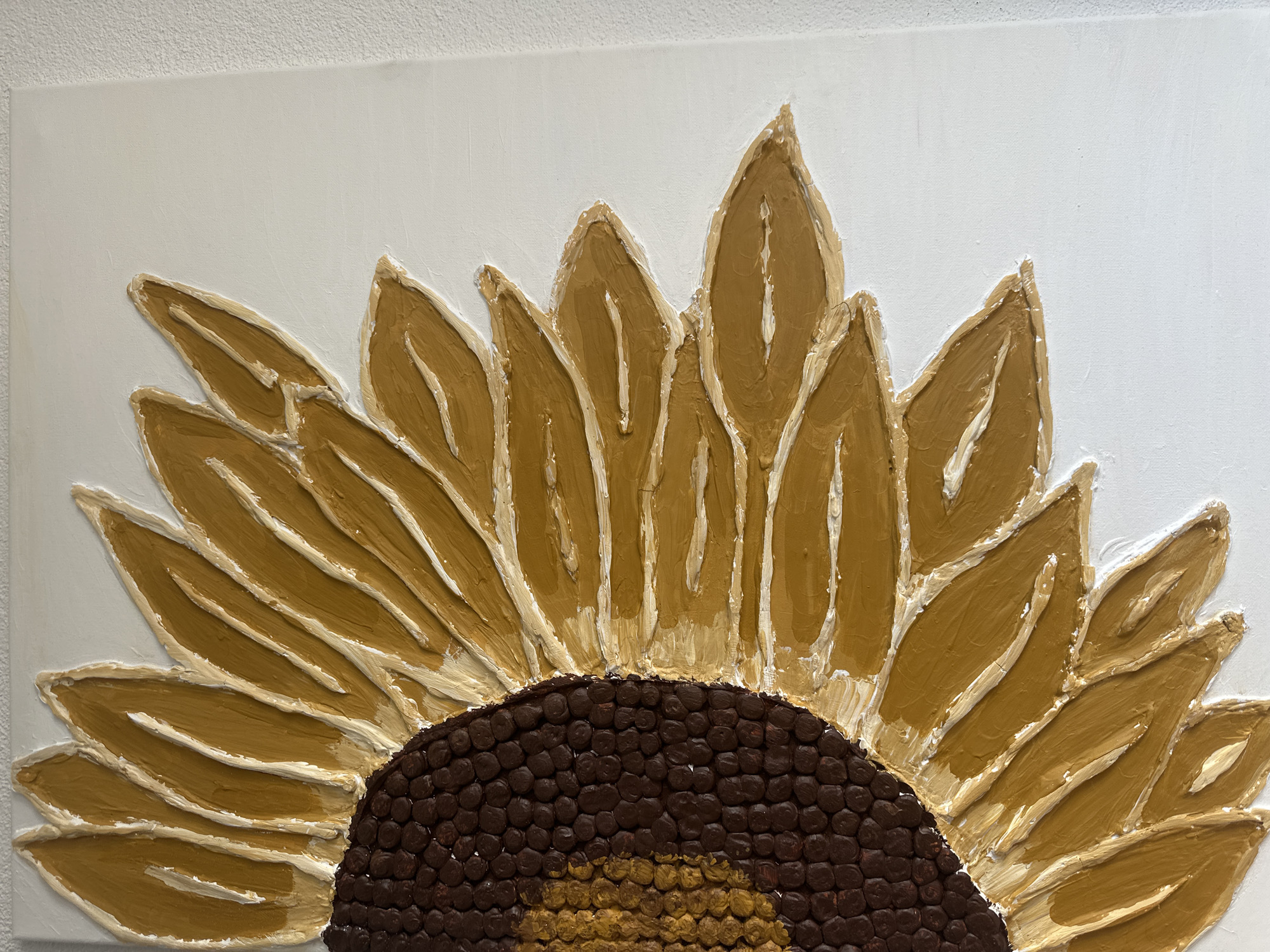 The image displays a close-up, textured painting of a sunflower's center and petals against a white background. The sunflower's center is depicted with dark brown circular shapes, resembling seeds, with a hint of golden yellow in the very center. The petals are rendered in a thick, impasto style, using shades of golden yellow and white, with visible brushstrokes and a raised, almost sculptural quality. The painting is mounted on a white canvas, and the background appears to be a textured wall. There is no identifiable location in the city of Günzburg, Germany, depicted in this image.