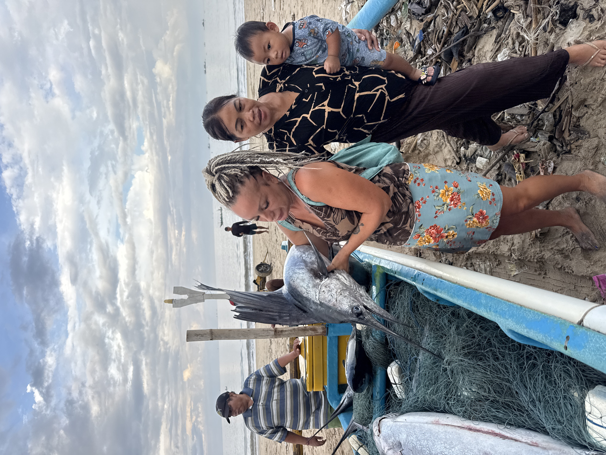 The image shows a group of people with a large fish on a beach in Kuta, Indonesia. A woman with blonde braided hair, wearing a floral dress, is examining the fish, which is a large marlin lying in a blue and yellow boat. A man in a striped shirt and baseball cap stands near the boat. Another woman stands behind the boat, holding a baby. The beach is sandy and there is some debris visible. The sky is cloudy.