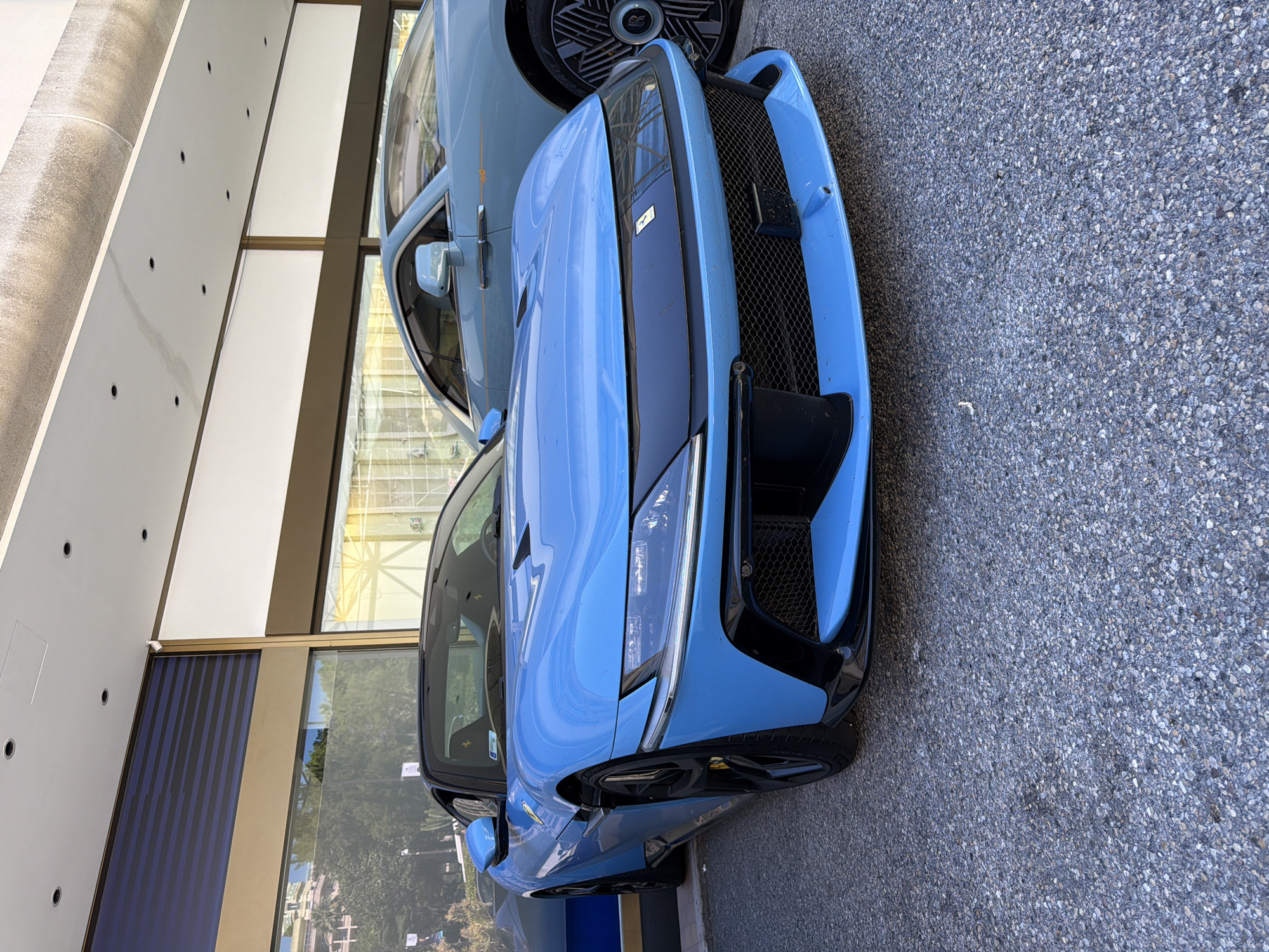 A bright blue Ferrari sports car is parked on an asphalt surface in front of a building with large glass windows and a beige and dark blue facade. The Ferrari's front end is prominent, featuring a black grille and sleek headlights. Behind the Ferrari, the rear half of a light blue Rolls-Royce car is visible. The background through the glass windows reveals trees and buildings. The scene is illuminated by daylight, casting some shadows.
