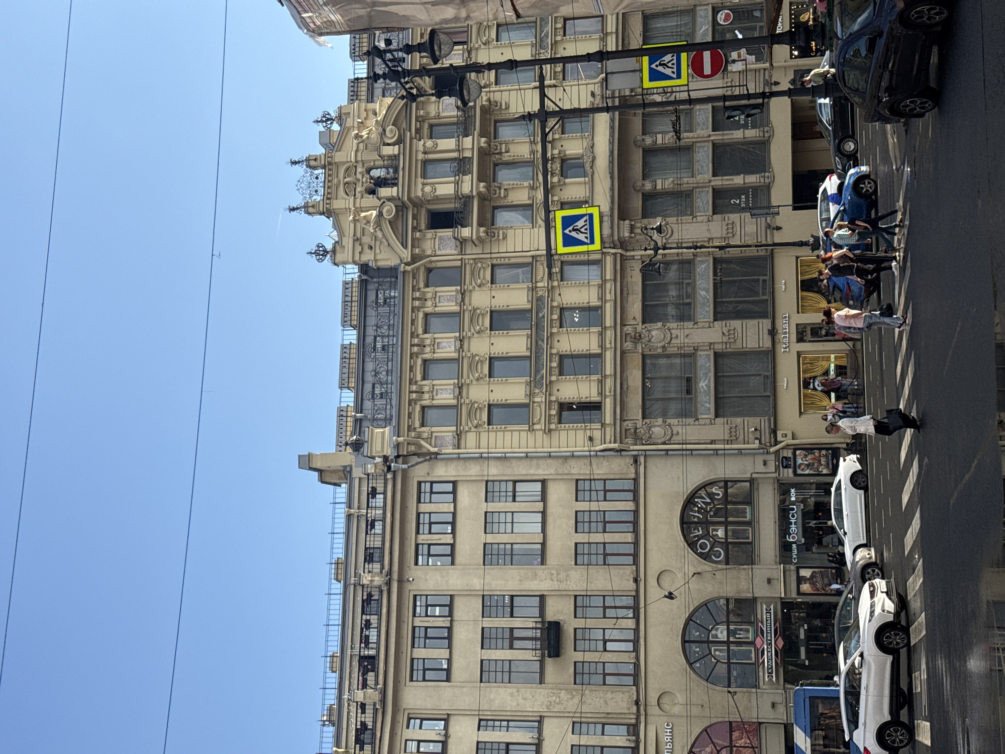 This image captures a street scene in Saint Petersburg, Russia, likely on Nevsky Prospect, given the architectural style and street signs. The foreground shows a wet asphalt road with a pedestrian crossing marked by white stripes. Several cars are visible, including a white SUV, a white sedan, a dark blue sedan, and a black sedan. Pedestrians are crossing the street at the designated crossing.
The midground features a row of historic buildings with ornate facades and multiple stories. The buildings are a pale yellow color and feature numerous windows with dark frames. Shops and businesses are located on the ground floor, indicated by signage such as "COLLINS," "ХУДОЖЕСТВЕННЫЙ," "СУШИ БЭНСИ," and "Italani." A blue bus is also partially visible on the left side of the image. Street lamps and traffic lights are present. Two pedestrian crossing signs, featuring a blue square with a white walking person symbol, are prominently displayed on poles. A red circular sign with a white horizontal bar, indicating "no entry" or a similar prohibition, is also visible.
The background is dominated by a clear, bright blue sky, with two thin dark horizontal lines that appear to be overhead wires. The architecture suggests the grandeur often associated with the city's historical center. The overall impression is a bustling urban environment on a sunny day, with the wet road indicating recent rain or a cleaning event.