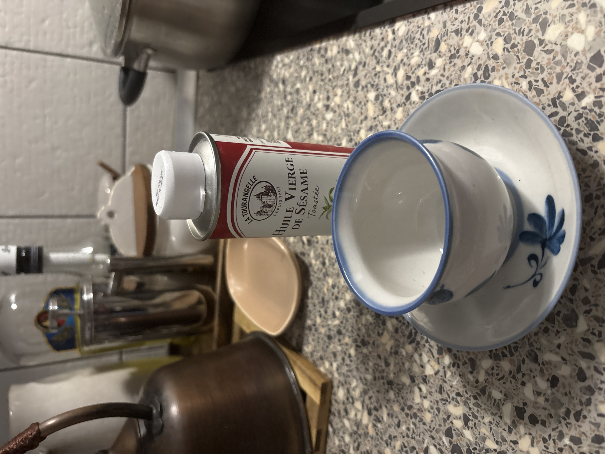 The image presents a countertop scene with various kitchen items. A La Tourangelle sesame oil can lies horizontally next to a white and blue teacup and saucer. Other objects in the background include a pot, bottles, and a wooden utensil holder with multiple items inside. The countertop has a speckled, mottled pattern. The location is not Saint Petersburg, Russia.