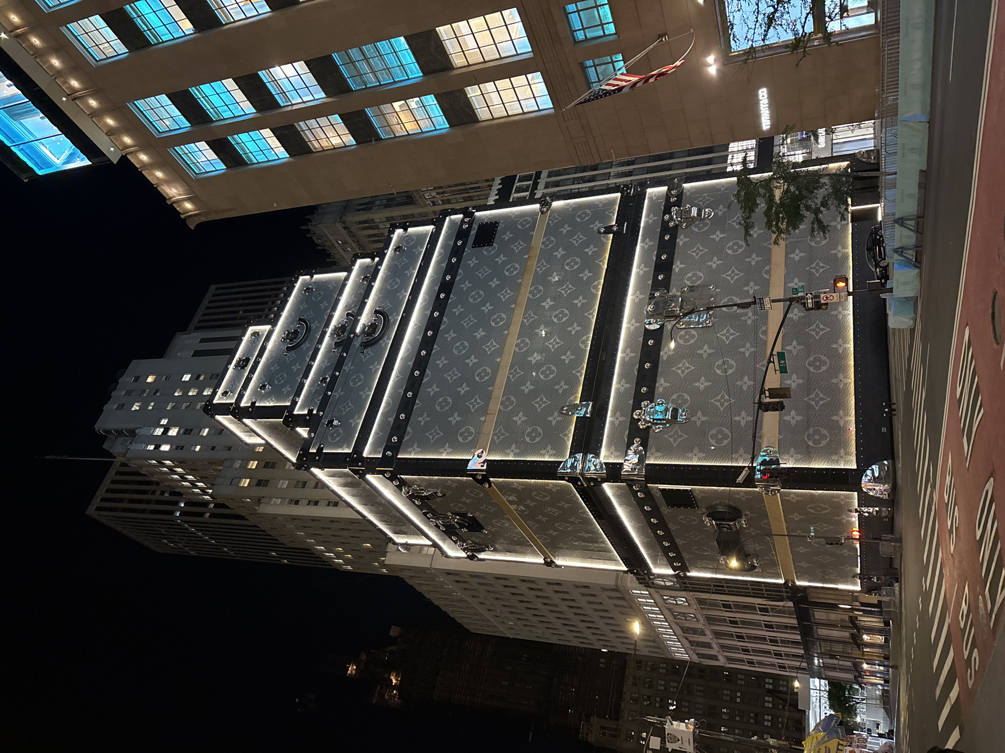 This nighttime image captures a vibrant urban street scene in New York City, USA, centered around a striking illuminated installation. The primary subject is a large, multi-tiered structure designed to resemble oversized Louis Vuitton trunks, covered with the brand's iconic monogram pattern and outlined by bright LED lights. This installation stands prominently along a city street.
Above and to the left of the trunk installation, a grand building with numerous windows, many glowing with a blue hue, dominates the upper part of the frame. The illuminated text "TIFFANY & CO." is visible on its facade, identifying the luxury retail establishment. An American flag hangs from this building.
To the right of the Louis Vuitton structure, a street is visible, complete with traffic lights and street markings. The text "BUS ONLY" is clearly painted on the road surface, indicating a dedicated lane for buses. A street sign displaying "5 Av" is also visible, suggesting the location is likely Fifth Avenue. In the background, other tall, illuminated city buildings rise under the dark night sky. Some trees with sparse foliage are also present in the urban setting.
The scene depicts an active commercial district at night, with the illuminated structures and street lights casting a glow. No specific human activities or individuals are clearly discernible, but the overall impression is one of a busy, upscale urban environment after dark. The weather appears clear with no visible precipitation.