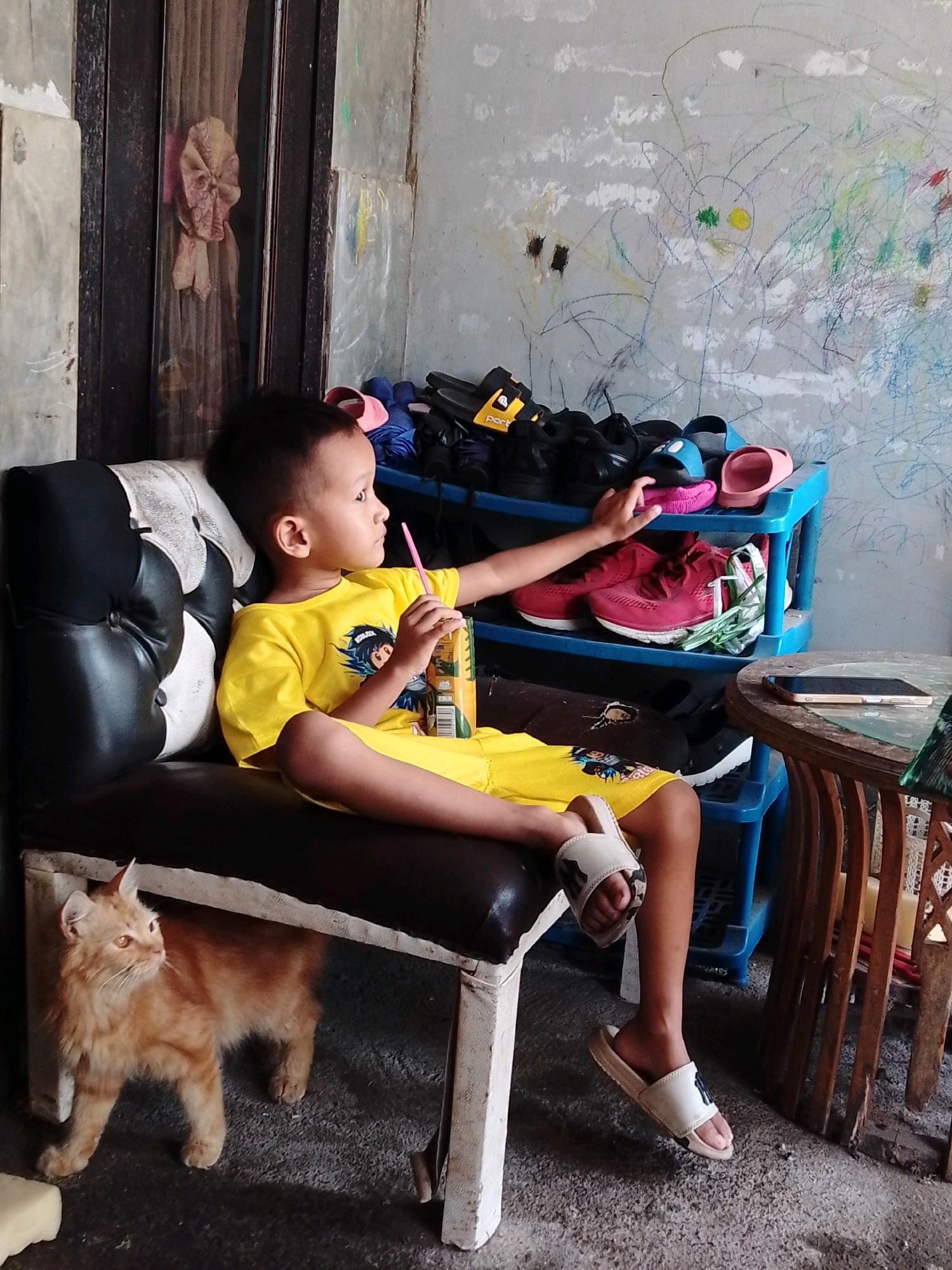 An Indonesian boy, appearing to be between 5 and 7 years old, sits on a dark cushioned bench with white trim. He is wearing a yellow t-shirt with a cartoon graphic and matching yellow shorts. He is holding a carton of juice with a straw in his right hand and is looking to his right. His feet are bare, but he is wearing white slip-on sandals with a black emblem. To his left, a ginger cat stands on the concrete floor, looking to the left. Behind the boy is a dark wooden door with a sheer curtain that has a pink and white patterned fabric tied to it. To the boy's right is a blue plastic shoe rack filled with various pairs of shoes and sandals, including pink slides, blue flip-flops, and red sneakers. Above the shoe rack, the concrete wall is covered in colorful crayon drawings. A round wooden table with a darker wood base is to the right of the shoe rack, with a smartphone resting on its surface. The overall environment suggests an outdoor or semi-outdoor space, possibly a home's exterior or a communal area in Cikal, Indonesia.