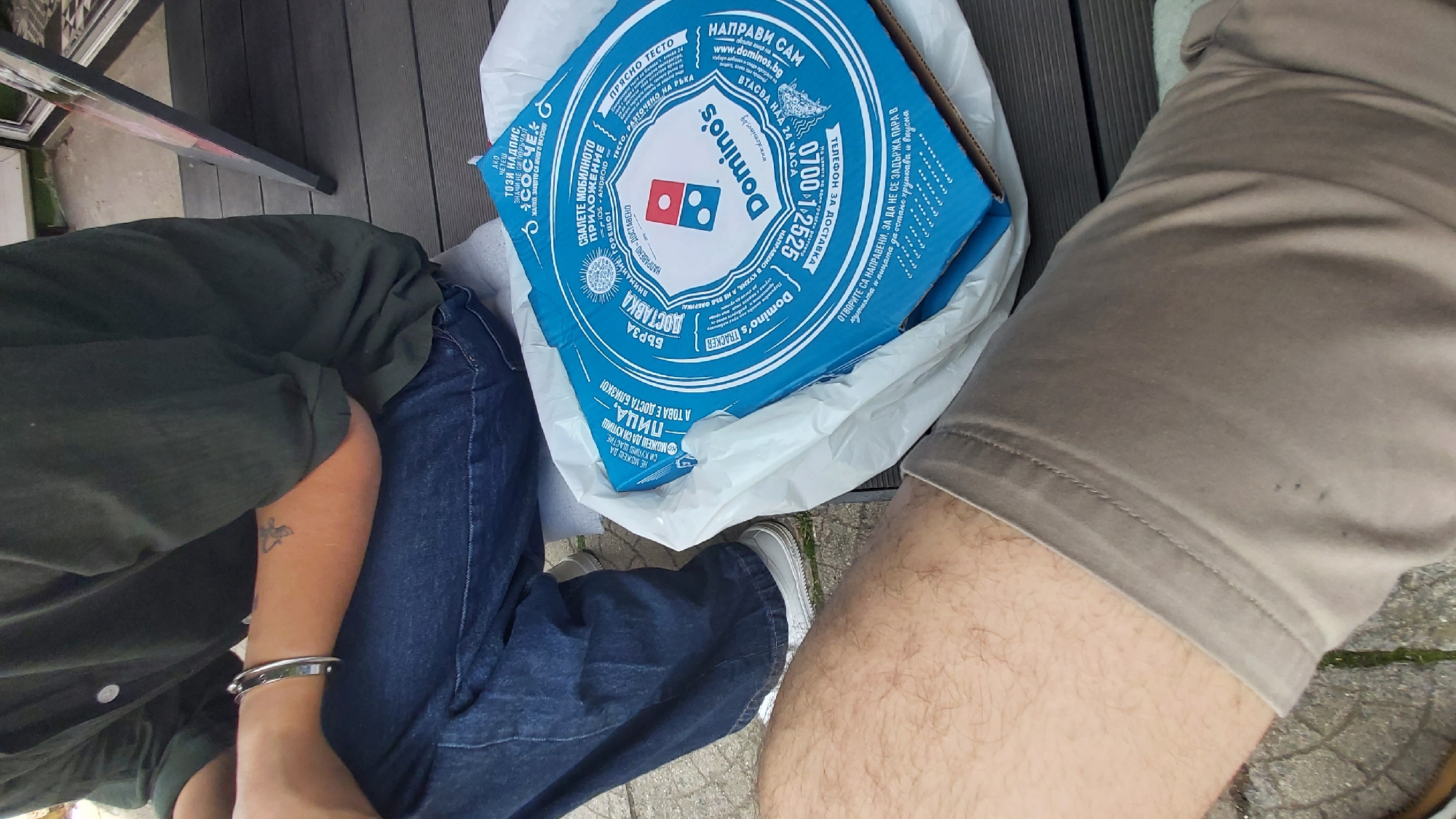 The image shows two people sitting outside, possibly in a public area in Sofia, Bulgaria, as evidenced by the Domino's pizza box which has Bulgarian text. One person is wearing a dark green shirt and jeans, with a silver bracelet and a tattoo on their left forearm. The other person is wearing khaki shorts. The Domino's pizza box is in a white plastic bag and rests between them on the ground. Part of a gray wooden floor is visible, suggesting an outdoor seating area. The ground is paved and has some grass growing.