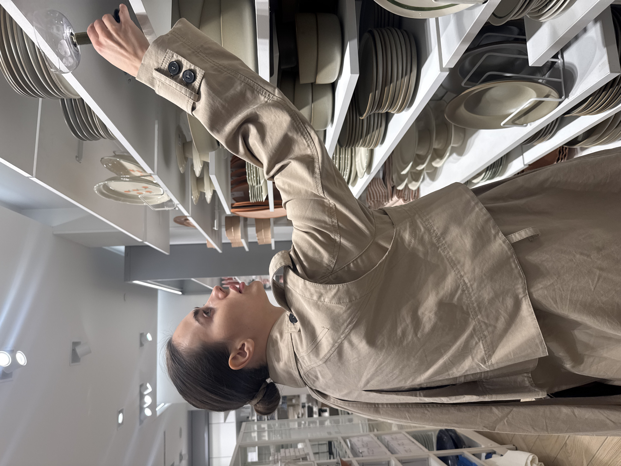 A woman wearing a tan trench coat is looking up at shelves filled with dishes in a store. The shelves are white and hold stacks of plates and bowls. The store appears to be a housewares or home goods store. The woman is reaching up with one arm. The background shows a retail environment with lighting fixtures and displays.