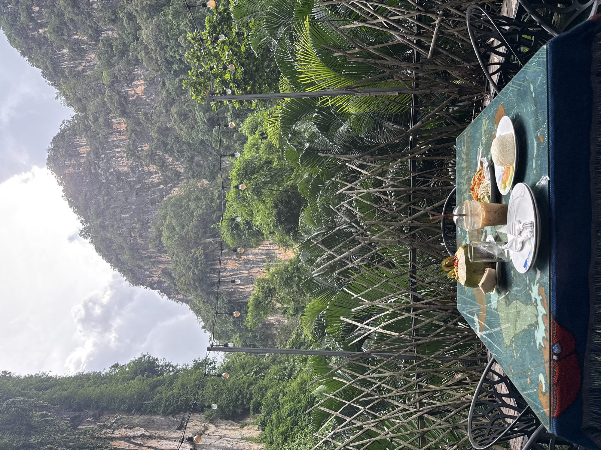 A table is set for a meal at an outdoor restaurant with a view of limestone cliffs and lush green foliage. The table is covered with a patterned tablecloth and set with plates of food, drinks, and cutlery. In the background, a lattice fence separates the dining area from the dense vegetation. String lights are hung overhead, and the scene is illuminated by natural light filtering through the clouds. The location appears to be in Chong Pli, Thailand, suggesting a natural and serene dining experience.