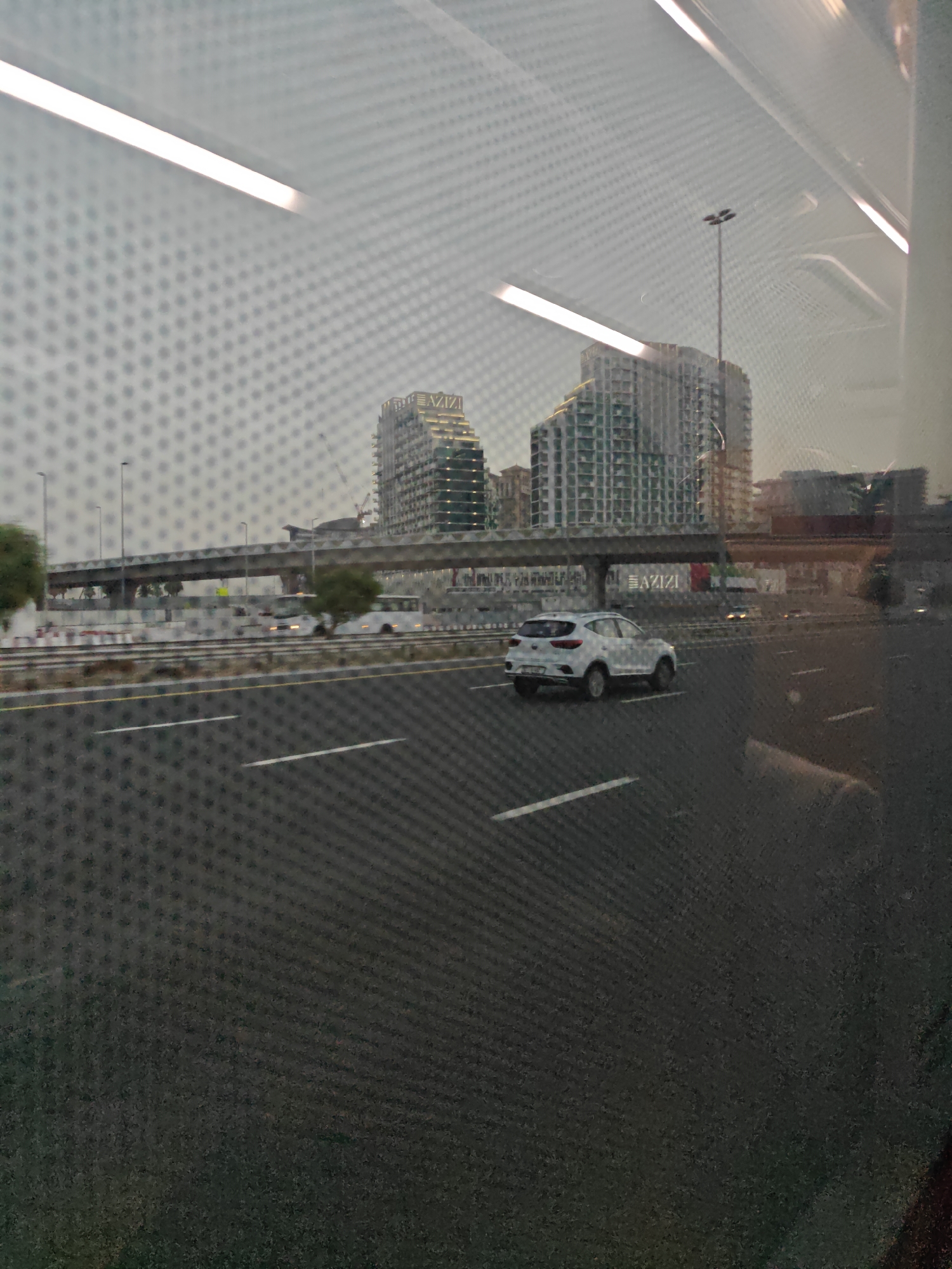 The image depicts a view from a vehicle, likely a bus or tram, looking out at an urban landscape in Dubai, United Arab Emirates. A multi-lane highway with white lane markings dominates the foreground, with a white SUV driving on the right side of the road. In the background, an elevated highway or metro line runs horizontally across the frame, supported by concrete pillars. Several modern, high-rise buildings with distinctive architectural designs are visible behind the elevated structure. One building in particular, on the right side of the frame, features the "AZIZI" logo prominently displayed. The sky is overcast, and the overall lighting suggests either early morning or late afternoon. The image is characterized by a grid-like pattern overlaid on the scene, indicative of viewing through a mesh or a patterned window.